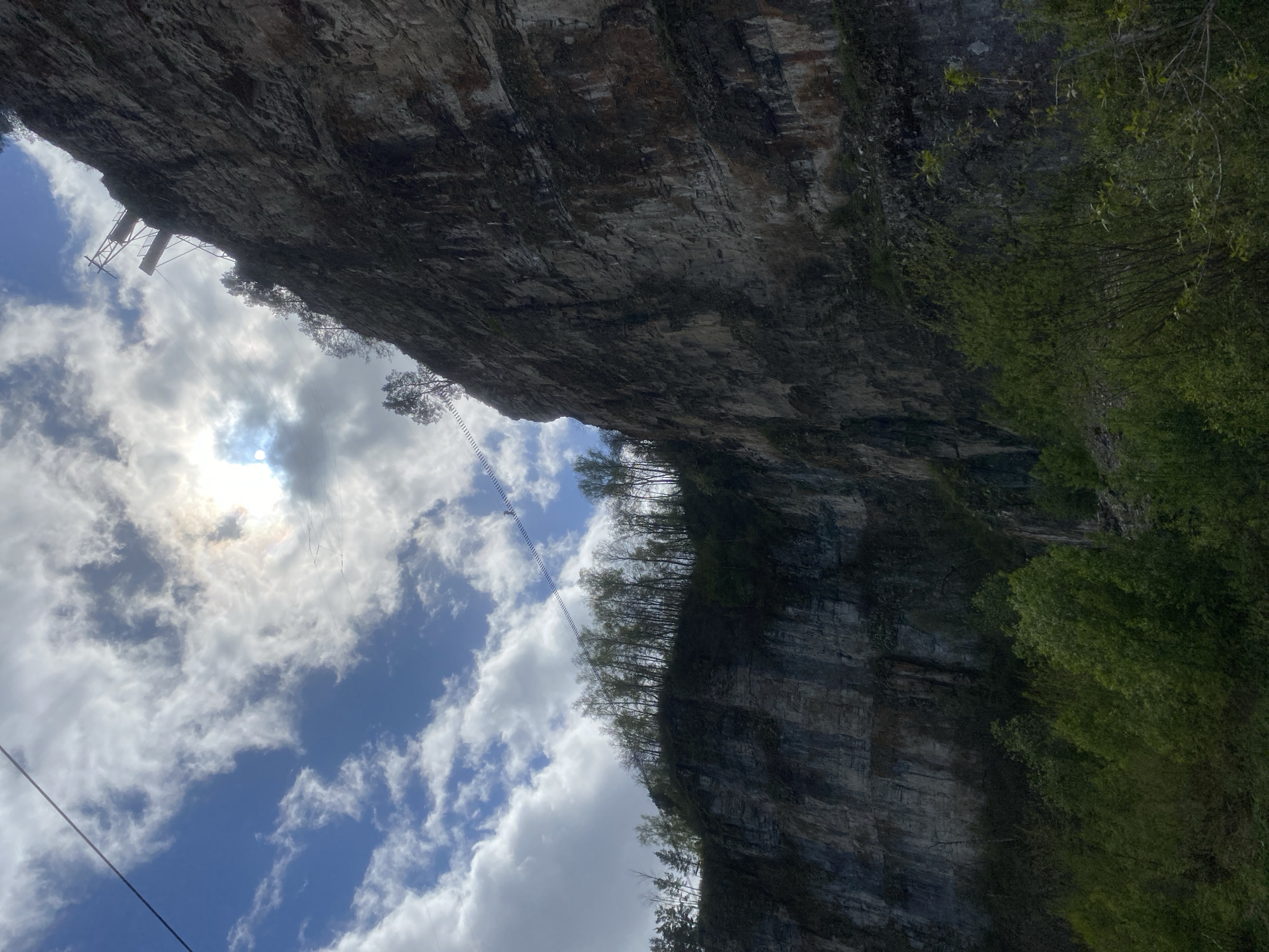 The image shows a view looking upwards towards a rock face under a sky of blue with scattered white clouds. On the upper-left side of the rock face is a metal tower structure connected to cables, which appear to run across the open space to the opposite rock face. Green vegetation grows along the edges of the rock face, indicating a natural environment. The photo orientation is tilted to the right. The location is Unknown, Russia.