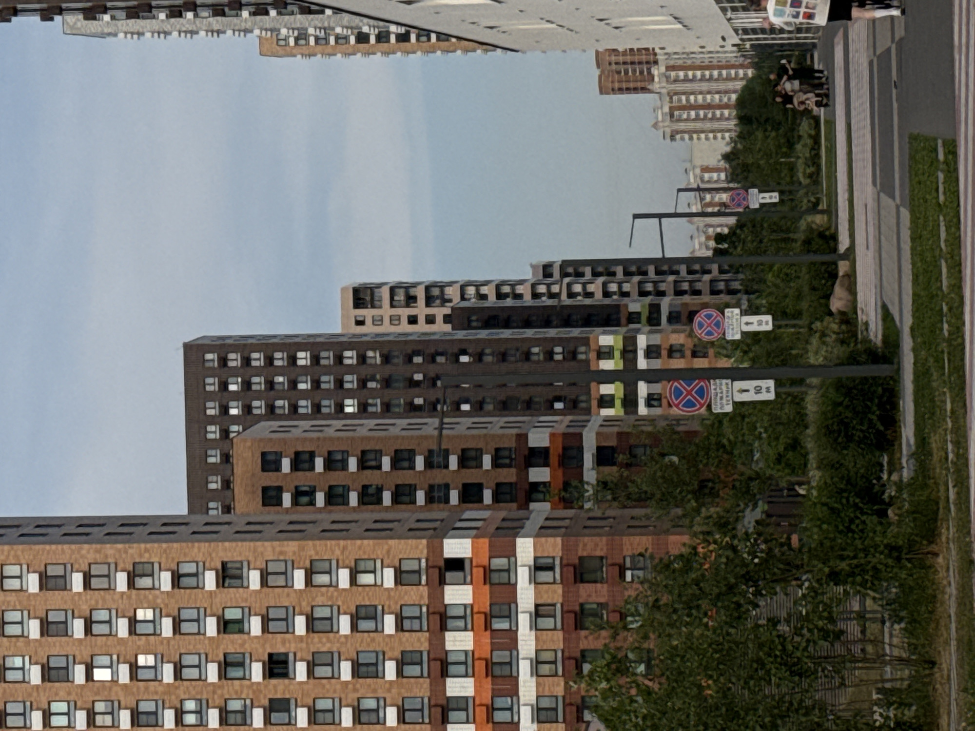The image shows a street scene in Moscow, Russia, with several tall residential buildings lining the street. The buildings are of modern design, with many windows and balconies. There are some trees and bushes along the sidewalk, and a few people can be seen walking on the street. There are also some traffic signs visible. The sky is overcast, suggesting it might be a cloudy day. The architecture of the buildings, particularly the style of the balconies and window frames, is characteristic of modern Russian urban development.
