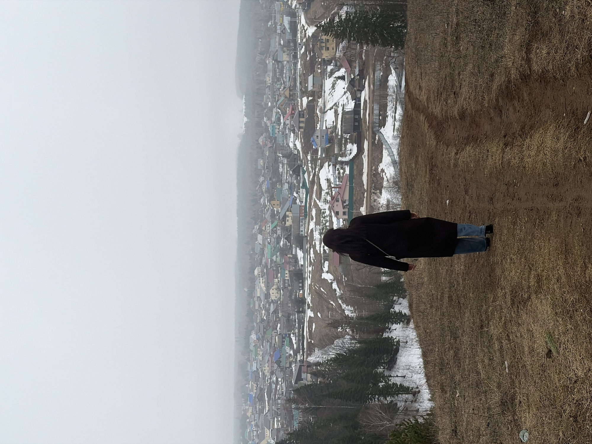 A person in a black coat and blue jeans stands on the edge of a grassy hill overlooking a village in a densely populated area in Ольховка, Russia. The village consists of small houses, and many have colored roofs. The ground has patches of snow. The sky is covered in fog.