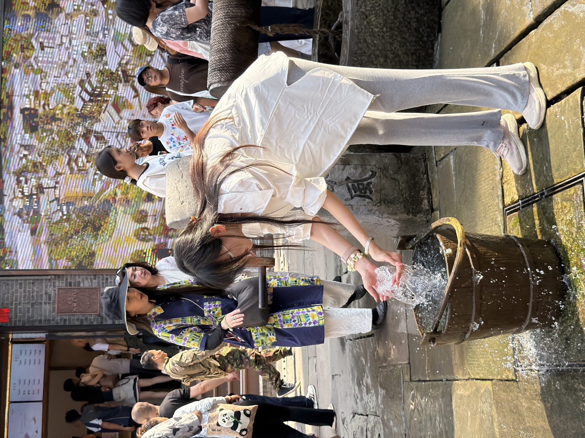 In Yuzhong District, China, on a sun-drenched cobblestone street, a young woman with long dark hair is captured in a moment of interaction with a weathered wooden bucket. She is dressed in a flowing white blouse and loose-fitting grey pants, her hands cupped to scoop water from the bucket, causing a splash. Two bracelets adorn her left wrist, one featuring small white beads and the other a silver chain. She is wearing pink athletic shoes. Behind her, a woman in a wide-brimmed hat and a vibrant, patterned kimono-style jacket stands with a black handbag. Other individuals, including a man in camouflage attire and a woman with a panda-themed tote bag, are visible in the background amidst a bustling urban setting, with a large digital screen displaying a colorful mural. The environment appears to be an outdoor market or historical street, with stone paving and architectural elements suggesting a traditional Chinese setting.