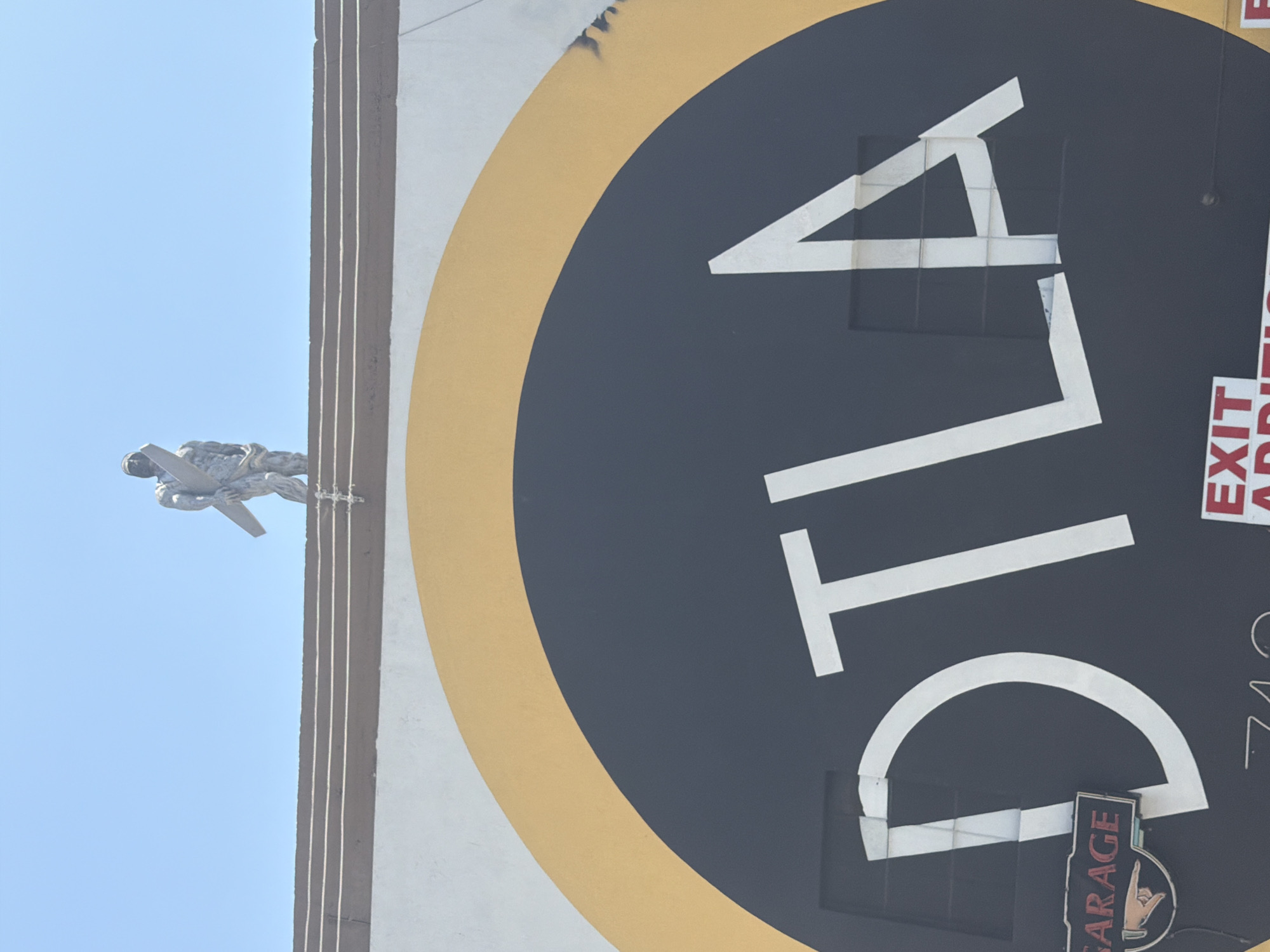 The image shows a large circular sign with the text "DLA" in white block letters against a dark background. A yellow stripe borders the dark circle. To the left of the sign, a statue of a man in mid-air, possibly a pilot or an aviator, is visible against a bright blue sky. The statue appears to be made of metal and is in a dynamic pose, with one arm extended forward, holding what looks like a wing or propeller.
The setting is outdoors, likely on a sunny day given the bright sky and clear lighting. The statue is positioned on the left side of the frame, appearing to be suspended or mounted on a structure that extends vertically. This structure is partially visible on the left, featuring dark, vertical lines that create a ribbed effect. The large sign occupies the majority of the right side of the image, with the letters "DLA" very prominent.
On the right edge of the sign, there is vertical text that reads "EXIT" in red and white letters. Below this, there is more text that is partially obscured but appears to include "APPRU." Further down, a small sign with the word "GARAGE" is visible, featuring a hand gesture that resembles the "shaka" sign. The overall scene suggests an urban environment, possibly near an airport or a building with a connection to aviation or commerce, given the "DLA" lettering which could be an acronym for an organization. The style of the statue and the sign suggest a possibly retro or artistic design.