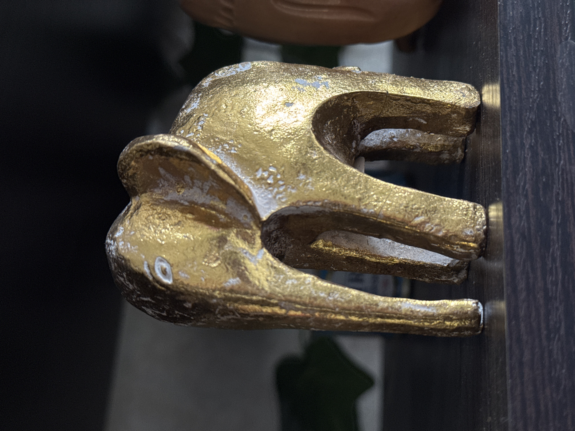The image shows a gold-painted elephant head-shaped handle or fixture attached to a dark-colored, possibly wooden, surface. The elephant's head has a stylized design with visible ears and a trunk. The gold paint appears to be weathered, with some areas showing wear or discoloration. The background is blurred, but suggests an indoor setting. No people are visible. The location of Novo-Nikol'skoe, Russia, is not directly discernable from the image.