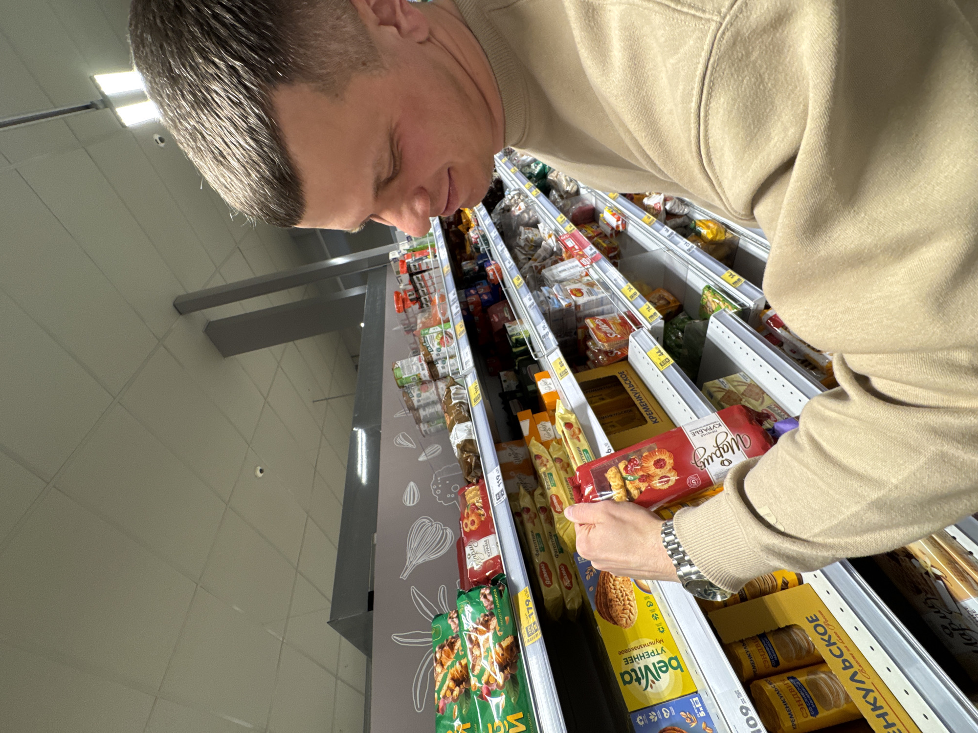 A man is seen selecting a package of "Mариз" cookies from a refrigerated display case, possibly in a store in the city of Горный, Russia. The case is filled with various food products including cookies and snacks. The man is wearing a light brown sweatshirt and a watch. The store features typical fluorescent ceiling lights and a tiled floor.
