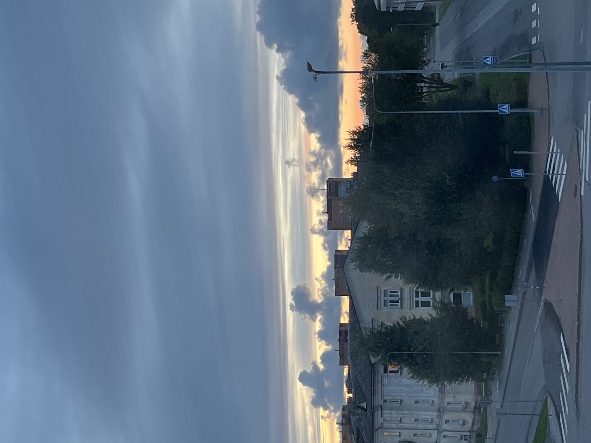 This is a medium-angle, eye-level, vertical shot of a street scene in Kohtla-Järve linn, Estonia, at dusk or dawn. The sky is overcast with dramatic clouds tinged with orange and yellow near the horizon, suggesting the sun is low. In the foreground, a street intersection is visible with white pedestrian crosswalks. To the left of the intersection, a sidewalk with traffic signs is present, including a blue sign with a white triangle and a figure walking. A tall, thin street lamp stands on the right side of the intersection. Behind the street, several trees are visible, with a large, multi-story building on the far left. Further back, more buildings, including a taller, modern one, are seen amongst the trees. The scene appears quiet and urban, with no people visible.