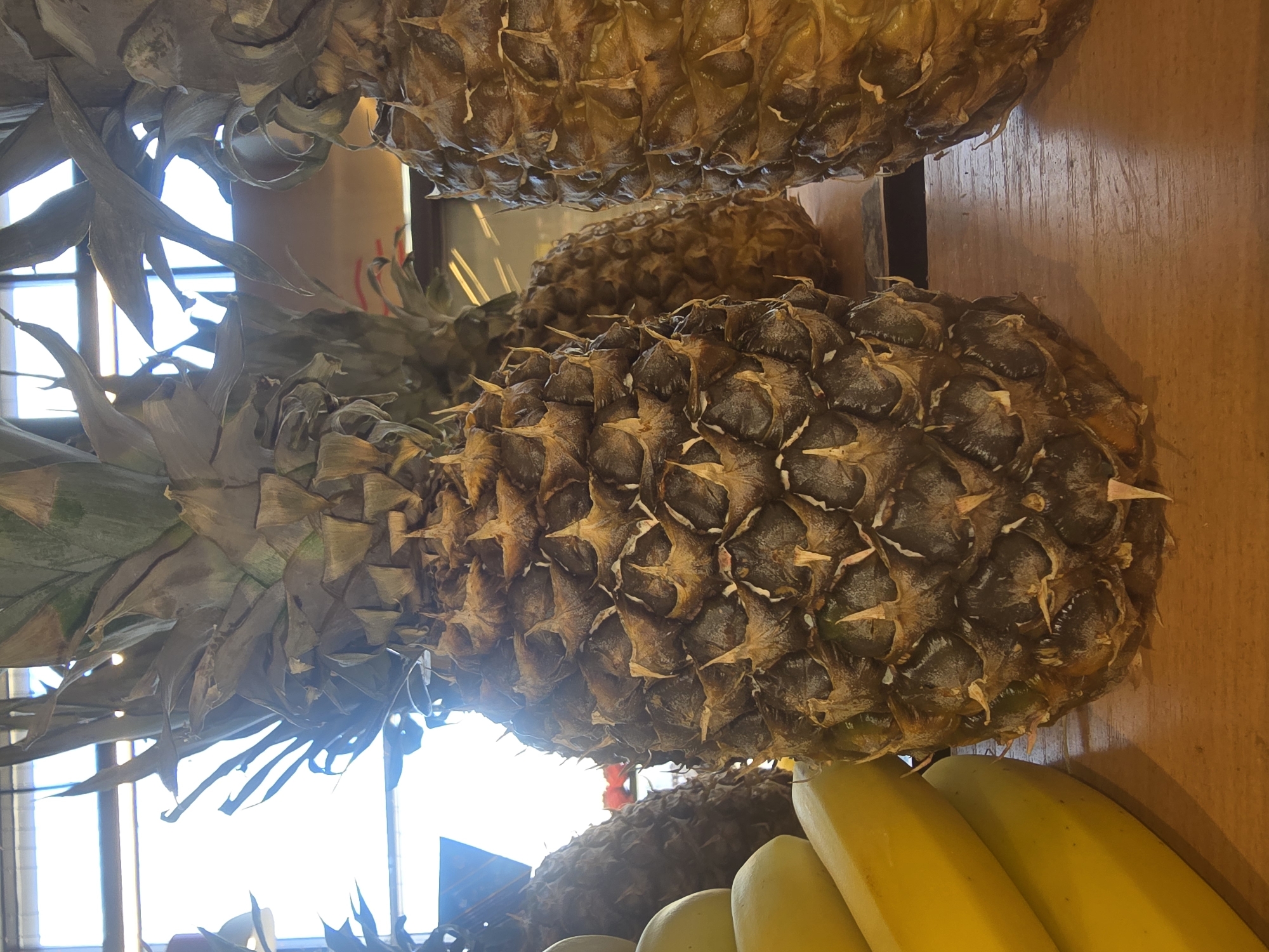 The image shows a close-up view of several pineapples and a bunch of bananas displayed on a wooden surface. The pineapples have rough, spiky exteriors and green leafy tops. The bananas are bright yellow. The background appears to be a window with some light filtering through. There is no discernible indication of the specific location in Ulan-Ude, Russia, nor any people or actions depicted. The scene is static and appears to be a still life arrangement of fruits.