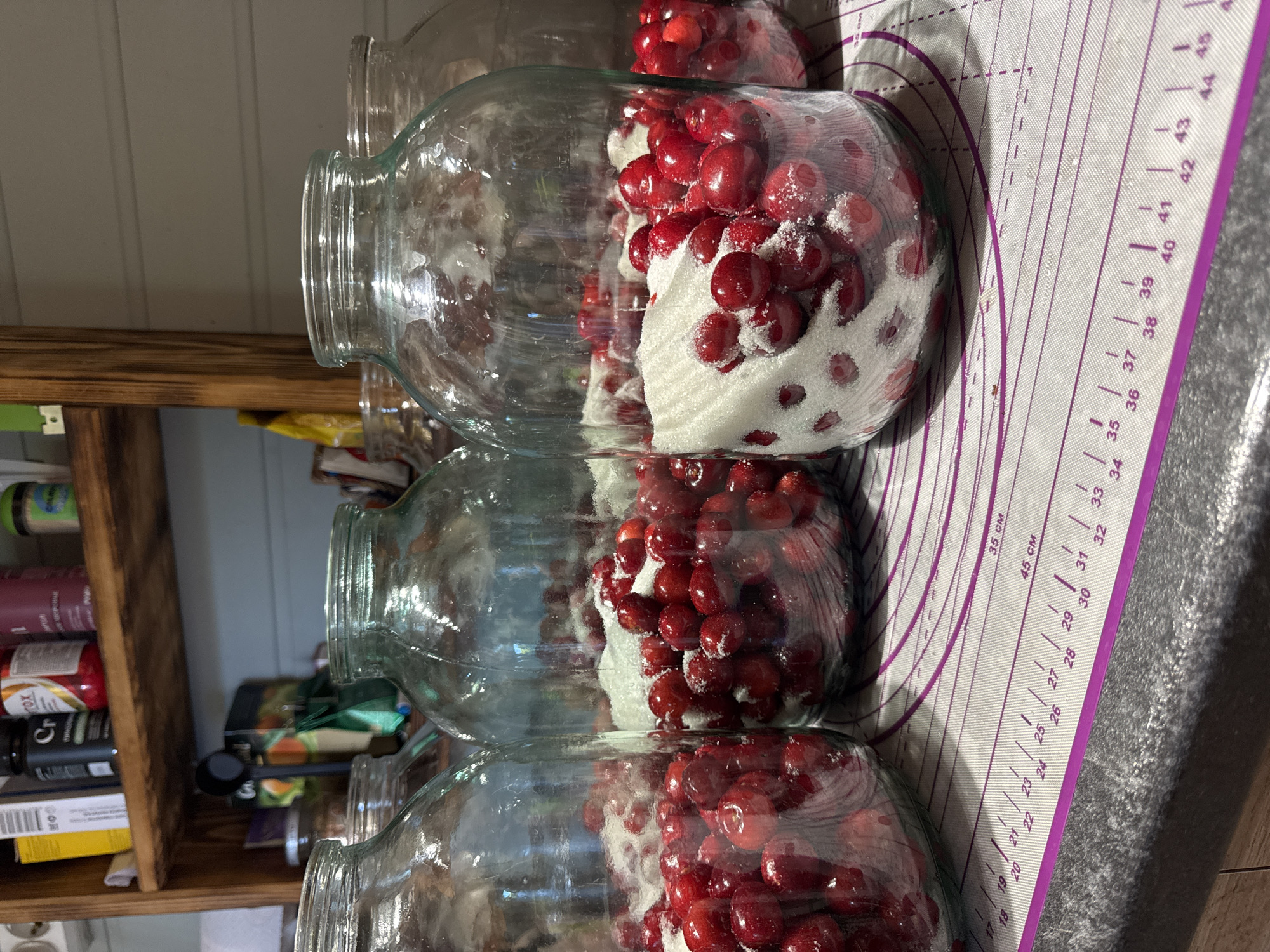 Three glass jars are stacked vertically, each containing fresh cherries and sugar. The jars are placed on a purple silicone mat with measurement markings. The contents of the jars are partially visible through the clear glass, showing a layered effect of red cherries interspersed with white sugar. The lighting suggests an indoor setting, possibly a kitchen, with natural light illuminating the scene. The overall atmosphere is one of preparation for food preservation or a sweet treat. Visible text on the mat includes measurement units and numbers in centimeters.