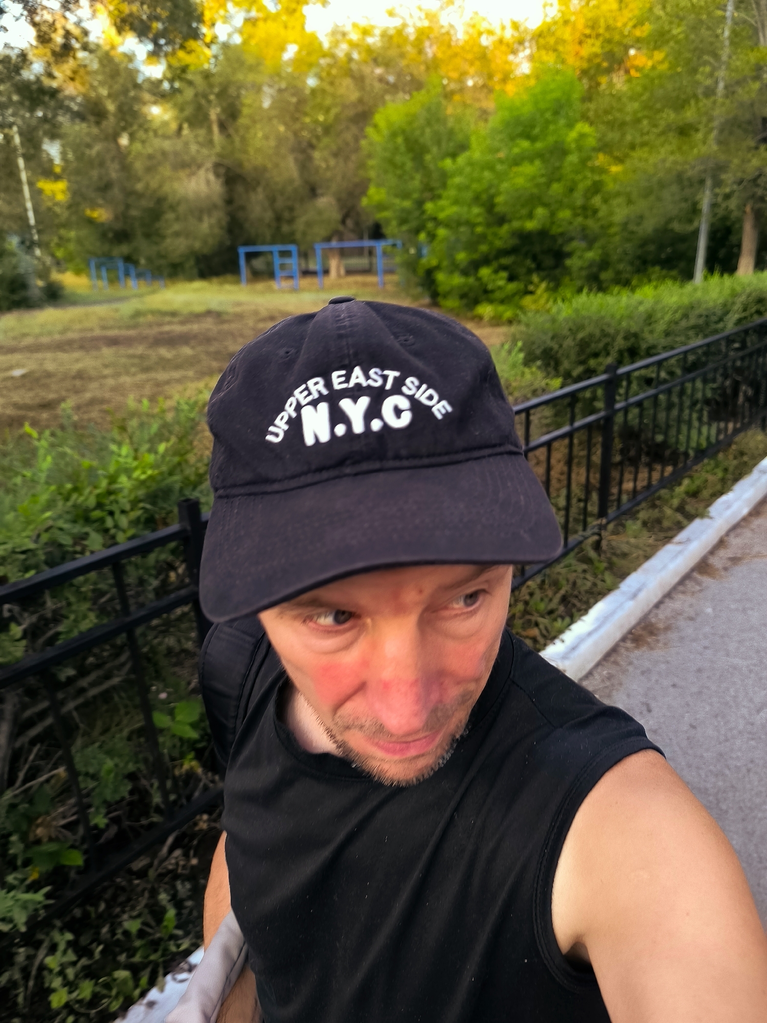 An eye-level, medium shot shows a man in a black sleeveless shirt and a black baseball cap with "UPPER EAST SIDE N.Y.C." embroidered in white. He is standing outdoors, facing slightly away from the camera to his right, with his right arm out of frame, presumably holding a camera for a selfie. His head and upper torso are in focus, with his face turned slightly upwards and to the right, and his mouth is slightly open. The background is blurred, showing trees with yellow and green foliage, a black metal fence, and a dirt path with some scattered greenery and blue structures in the distance. The lighting suggests it is either early morning or late afternoon. The location is described as the city of Qarağandı city administration, Kazakhstan, although this specific context is not visually discernible from the image itself.