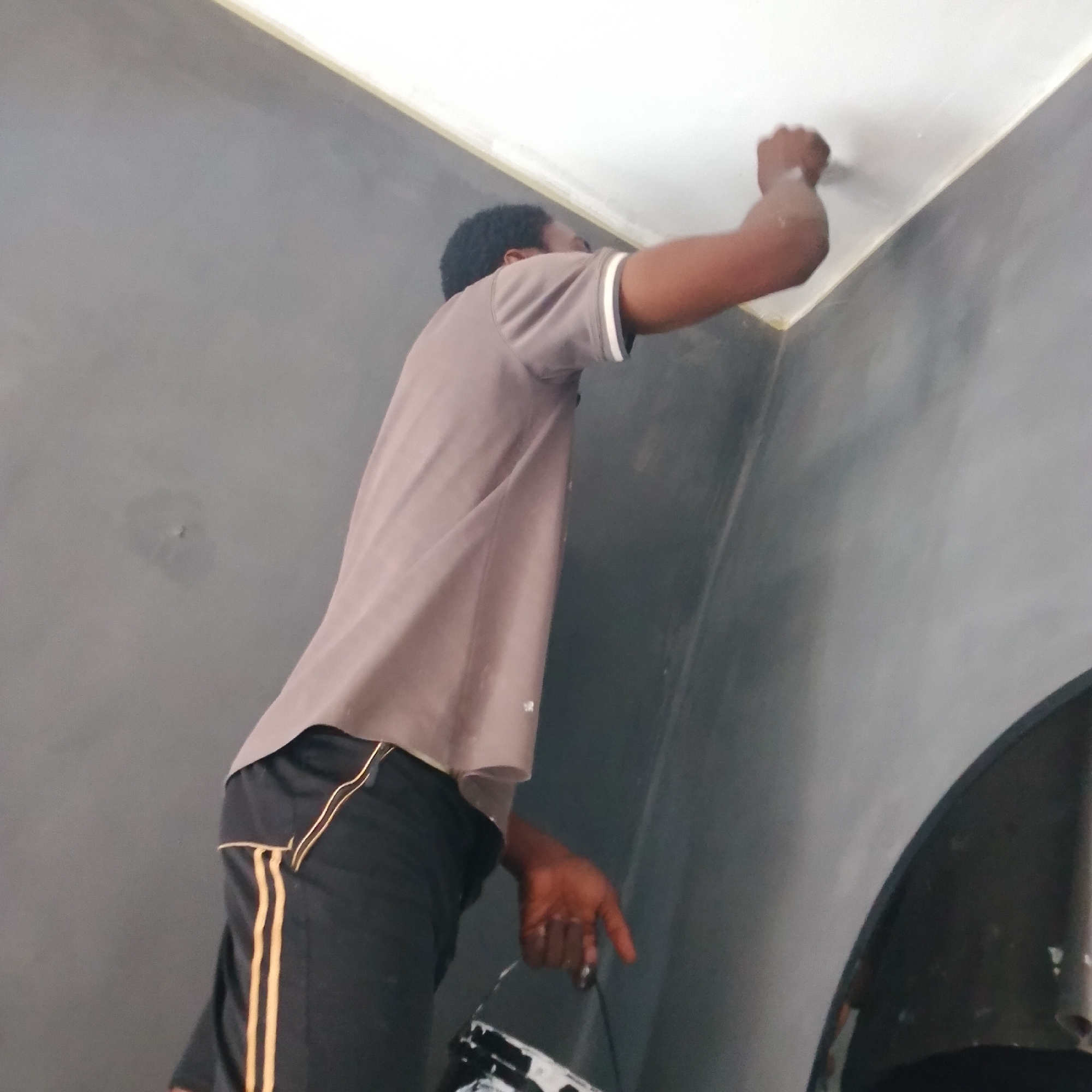 In Ado, Nigeria, a person is seen painting the ceiling of a room. The person is wearing a light brown t-shirt with white and grey stripes on the sleeves, and black shorts with yellow stripes. They are standing on an unseen object to reach the ceiling. Their right arm is raised, applying paint to the ceiling. In their left hand, they are holding a white object, possibly a paint roller or a tool. The walls of the room are painted a dark grey color. The ceiling appears to be white and is being painted with a light yellow trim along the edge where it meets the wall. The lighting suggests it is daytime, possibly indoors with some ambient light.