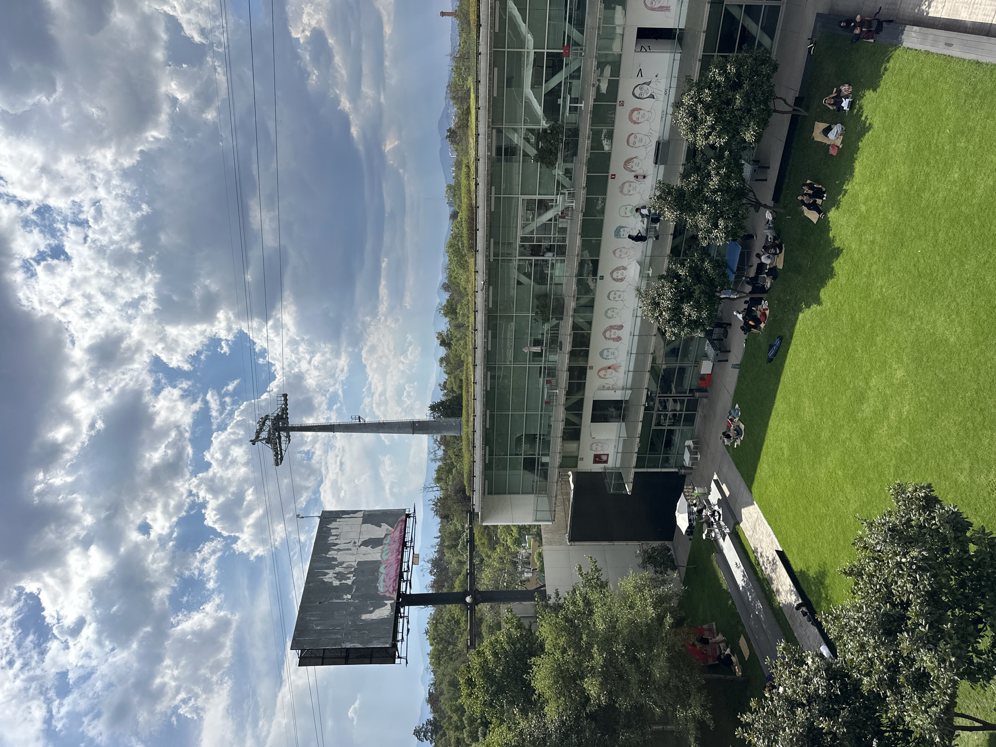 The image shows an aerial view of an outdoor space adjacent to a modern glass building, likely taken in Mexico City, Mexico. The foreground features a green lawn where several groups of people are sitting and reclining. Some individuals appear to be reading or working on laptops. Adjacent to the lawn is a walkway with people strolling and gathering near tables under a white umbrella. The building's facade is adorned with stylized drawings of faces. In the distance, a large billboard is visible, along with an electrical transmission tower. The sky is filled with puffy white clouds. The overall setting suggests a campus or corporate park environment.