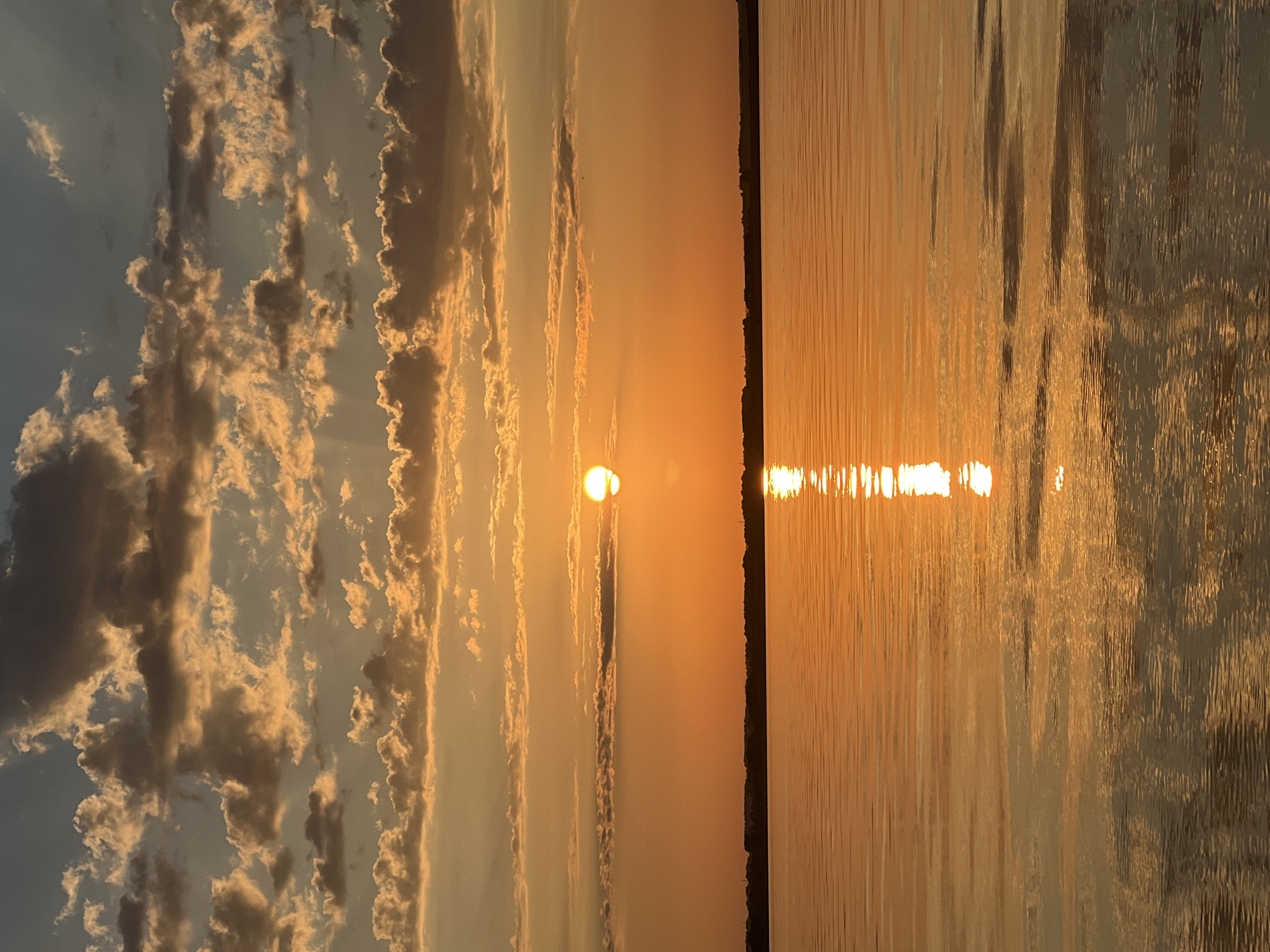 A sunset over the Ob River near Novopichugovo, Russia, is captured in a vertical, full-frame photograph. The sun, a bright, white disc, is low on the horizon, partially obscured by a thin band of clouds. Its rays cast a warm, orange glow across the sky, illuminating scattered, textured clouds ranging from pale yellow to deep grey. The water of the Ob River is calm, reflecting the vibrant colors of the sky and the sun's light in a shimmering path. A dark, silhouetted treeline marks the far bank of the river, with faint suggestions of distant structures. The overall impression is one of serene natural beauty.