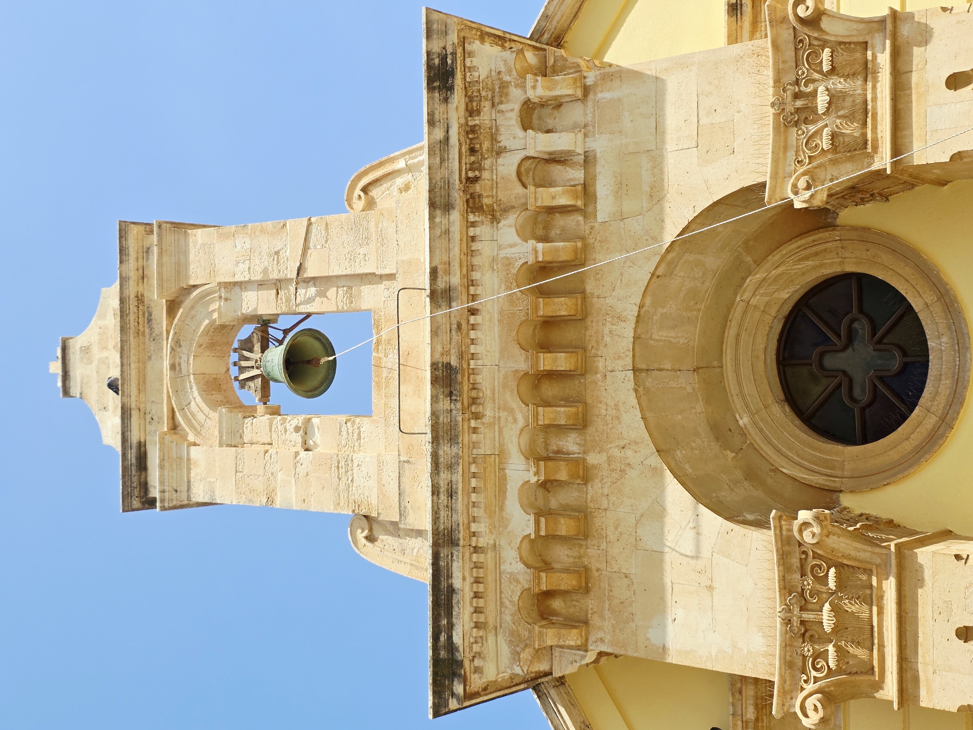 The image shows a bell tower of a church in Heraklion, Greece. The bell tower is made of stone and has a bell inside. The sky is blue and clear. The church appears to be old and historic. The image is taken from a low angle, looking up at the bell tower. There is a string hanging down from the bell. The architectural details suggest it may be a Baroque or Renaissance style.
