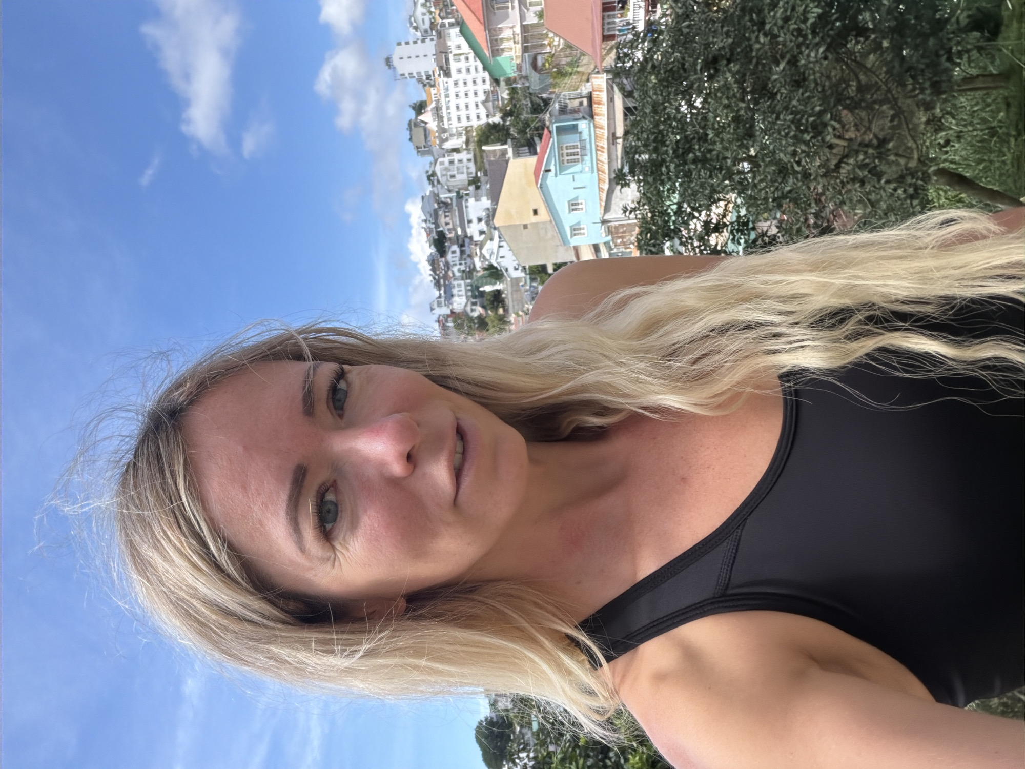A woman with blonde hair is taking a selfie outdoors, with a city skyline in the background. She is wearing a black tank top. The city appears to be Da Lat, Vietnam, characterized by its hilly terrain and densely packed houses with colorful roofs. The sky is blue with some scattered clouds. Trees are visible in the foreground.