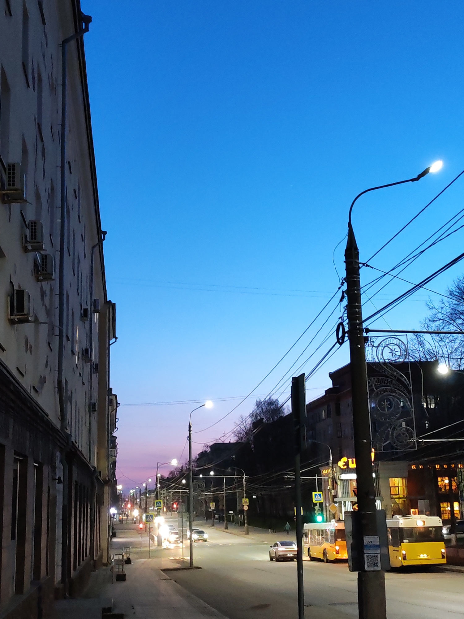 This image captures a vibrant urban street scene in Izhevsk, Russia, during twilight. The sky transitions from a deep, clear blue overhead to a softer, purplish-pink hue towards the horizon, indicating either late dusk or early dawn.
On the left, a tall, multi-story building with a light-colored facade dominates the foreground, featuring numerous windows and external air conditioning units along its side. A drainpipe runs vertically down its exterior. The sidewalk alongside this building is visible but mostly empty.
To the right, a busy street stretches into the distance, lined with streetlights that illuminate the scene with bright white and yellow light. Utility poles laden with numerous power and trolleybus wires crisscross the frame, adding to the urban density. Several vehicles, including cars with their headlights on, are visible on the road. In the mid-ground, two distinctive yellow buses, possibly trolleybuses given the overhead wires, are either stopped or moving slowly near a pedestrian crossing. A pedestrian can be seen walking on the sidewalk further down the street on the right.
Buildings in the background on the right have illuminated windows, suggesting commercial establishments. One building features an ornate metal decorative element or gate. Visible text includes "Евро" on a building sign in the mid-ground and a sticker on a utility pole with "15Я" and a QR code, likely providing route or stop information. The overall impression is that of a bustling city street transitioning from day to night, with the artificial lights creating a warm contrast against the cool twilight sky. The bare trees in the distance suggest it might be late autumn or winter.