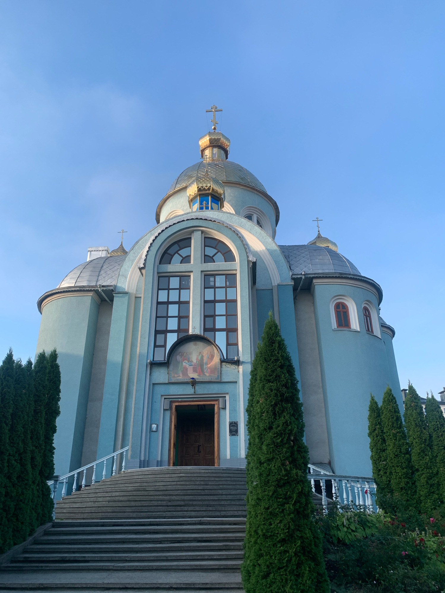 The image shows the exterior of the Church of the Blessed Virgin Mary in Kolomyia, Ukraine, on a clear day with a pale blue sky. The church features a prominent blue facade, a large central dome topped with a golden cross, and smaller domes on either side. A grand stone staircase with white railings leads up to the main entrance, which is a large wooden door. Above the entrance is a large arched window with a cross-shaped mullion and a religious fresco. Tall, dark green coniferous trees flank the staircase and the building. The overall scene is one of architectural grandeur and religious solemnity.