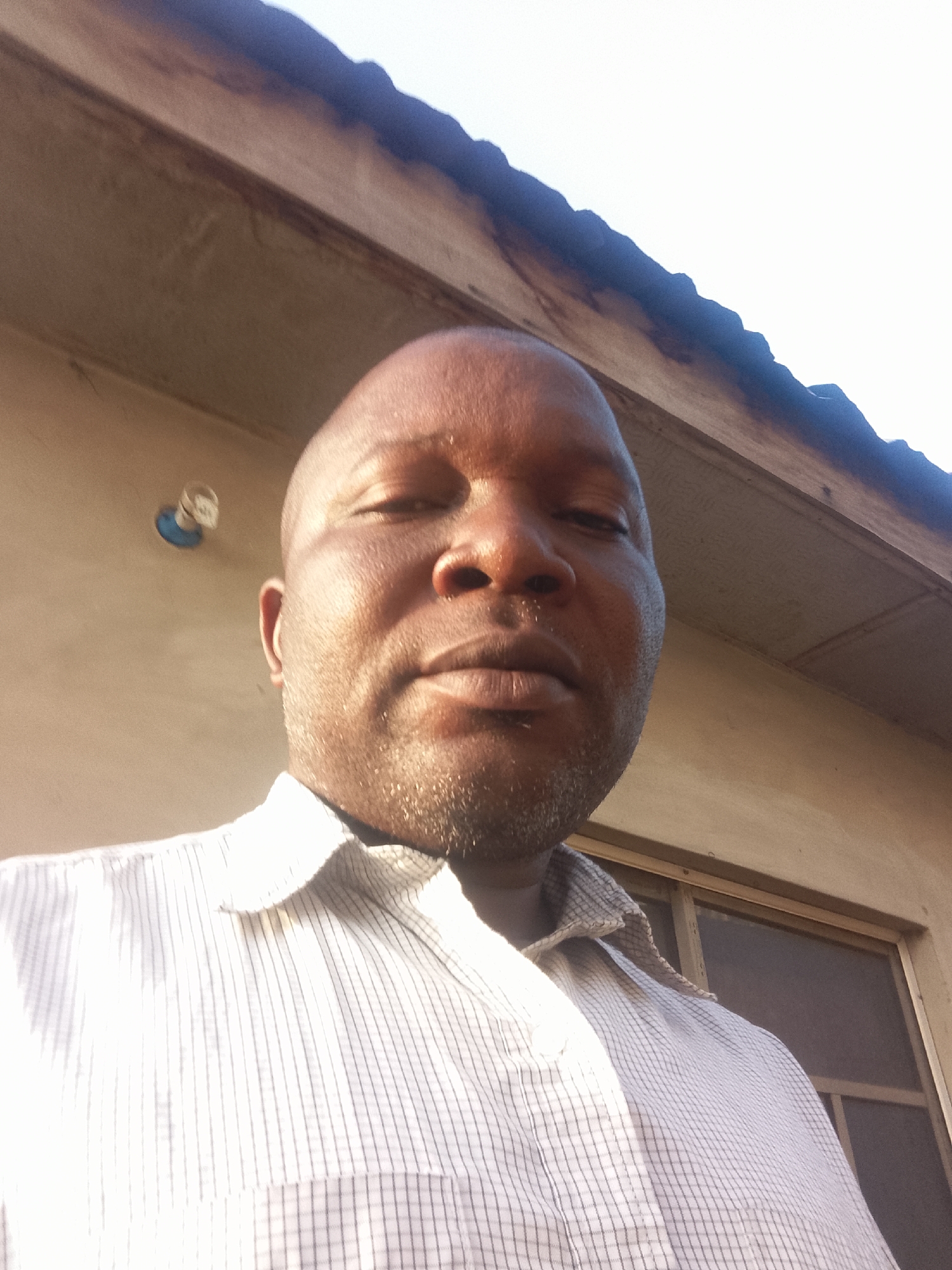 The image shows a close-up selfie of a bald, dark-skinned man wearing a white striped shirt. He appears to be standing outdoors near a building with a window and a light fixture. The specific location within the city of Unknown, Unknown, cannot be determined from the image.