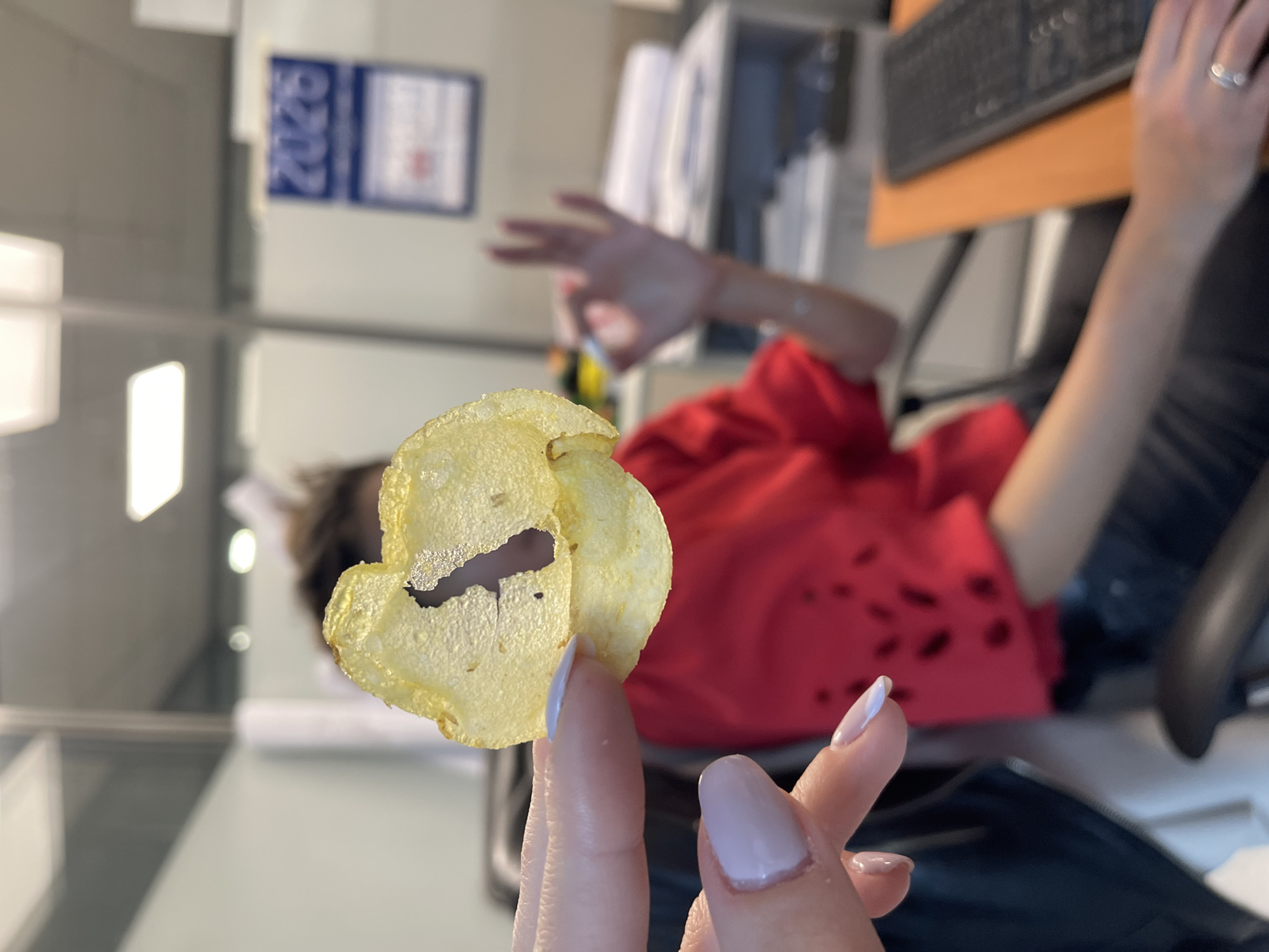 The image, captured from a first-person perspective and appearing rotated approximately 90 degrees clockwise, depicts a casual moment in an indoor office setting. In the immediate foreground, a hand with light pink-toned, almond-shaped manicured nails holds a single, light yellow potato chip. The chip is thin and appears crispy, with a noticeable hole in its center.
In the blurred background, another person is visible, positioned as if seated at a desk but appearing to lie down due to the image's orientation. This individual wears a red top, possibly a t-shirt or hoodie, which features a distressed or patterned texture. One arm is raised and extended towards the left of the frame, with fingers spread, while the other hand rests on a light brown wooden desk next to a black computer keyboard. A silver ring is visible on the ring finger of this resting hand. The person's face is out of focus and largely obscured.
The office environment is further indicated by elements such as a black office chair, other indistinct office furniture, and wall-mounted documents or posters. A blue document on the back wall clearly displays the numbers "2020". The overall activity suggests a snack break or a moment of leisure during work. No specific time of day or weather is discernible. The general setting is an office, with the provided context placing this location in Sofia, Bulgaria.
