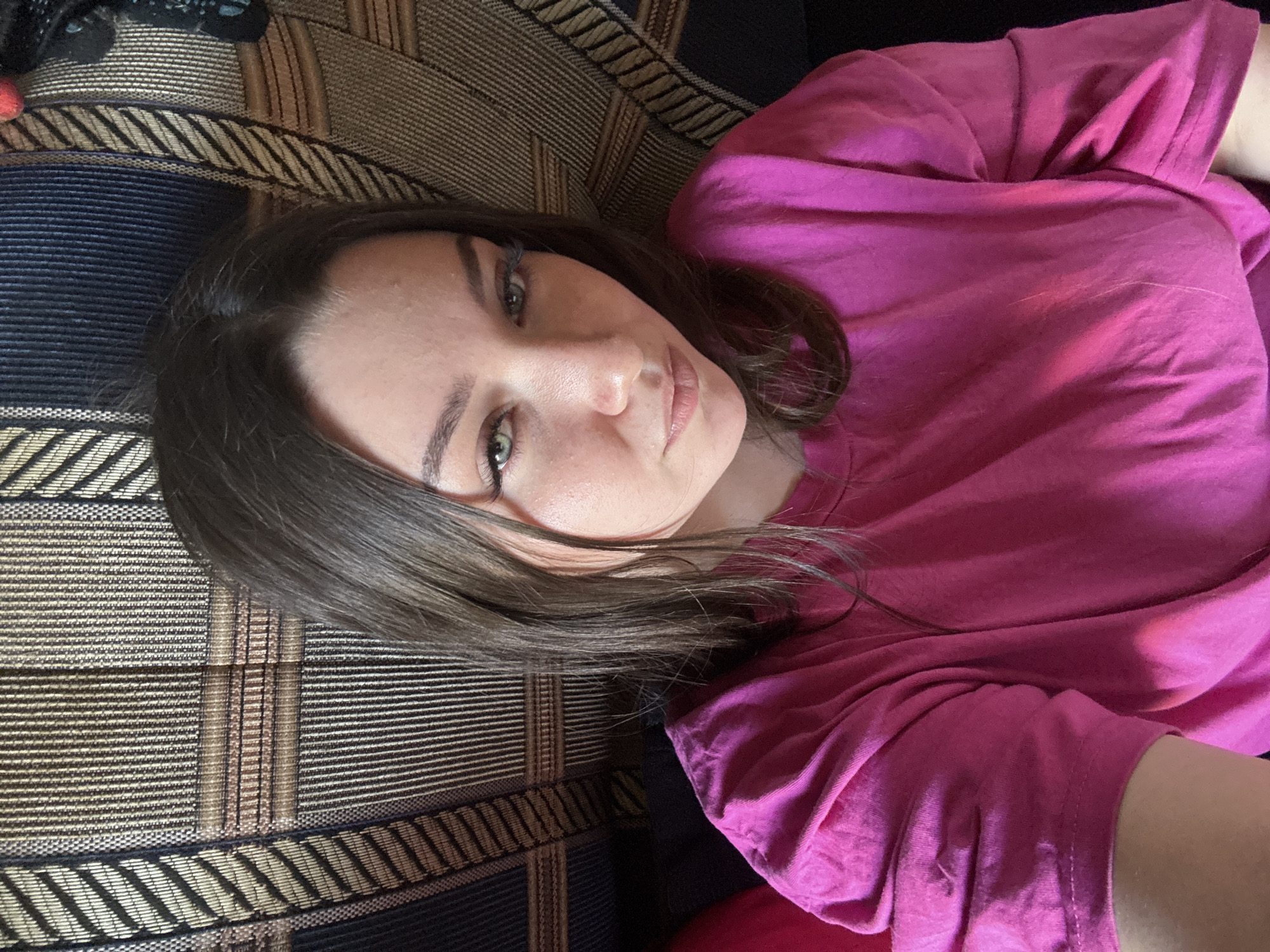 A close-up, top-down, portrait of a young woman is captured on a couch. She is lying on her side, facing the camera, with her head resting on a patterned sofa. She has medium-length, dark brown hair that falls around her face and shoulders. Her eyes are a striking green, and she is looking directly at the camera with a neutral expression. She is wearing a bright pink t-shirt, which is slightly creased. The sofa has a dark background with a woven pattern of thin horizontal lines in black, gold, and beige, with wider bands of blue and gold running vertically in sections. The lighting suggests it is daytime, and the scene appears to be indoors, likely in a home. There are no visible signs of text in the image. The overall mood is relaxed and casual.