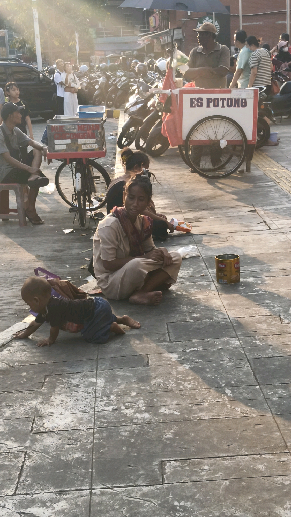 The image captures a street scene in West Jakarta, Indonesia, likely in the afternoon given the warm lighting and shadows. In the foreground, a young child is crawling on a textured concrete sidewalk. Slightly behind and to the right, a woman is sitting cross-legged on the ground, looking to her left. She is wearing a light-colored top and a patterned scarf around her neck. Next to her, a metal can is placed on the ground.
In the middle ground, a food cart with the text "ES POTONG" is visible, indicating it sells ice. A man wearing a cap is standing near the cart, facing left. Behind the cart, numerous motorcycles are parked, suggesting a busy area. Further back, several people are walking or standing, and some buildings and other structures are discernible. The overall environment suggests an outdoor public space within the city, possibly a market area or a busy street.