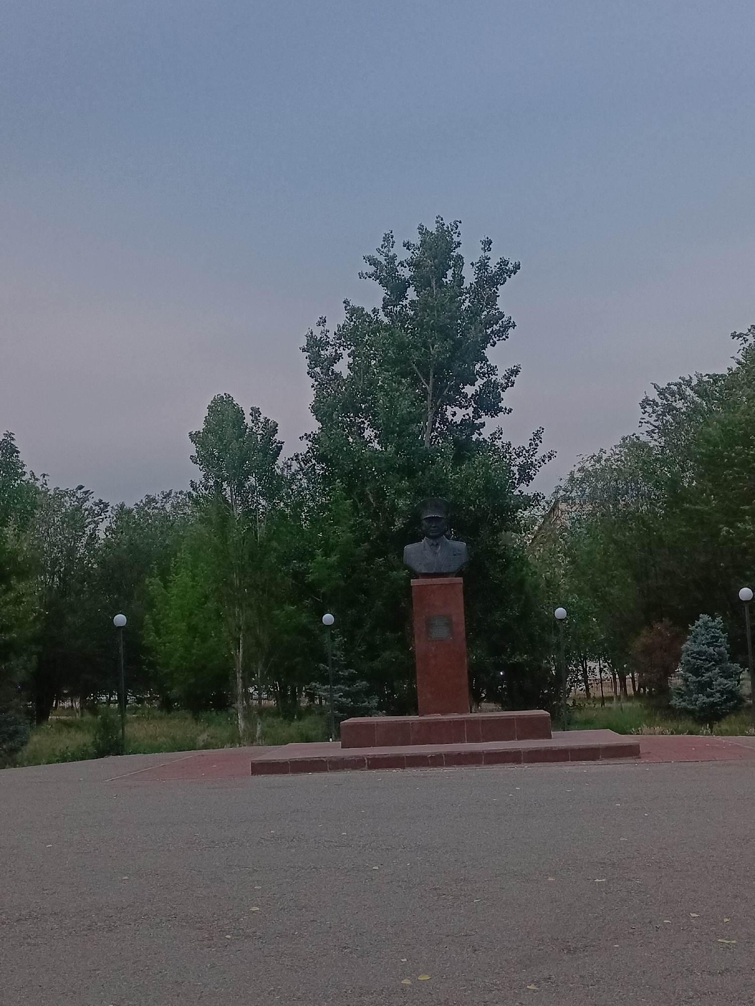 The image depicts a bust of a man in military uniform, wearing a cap, placed on a rectangular red stone pedestal. The pedestal is situated on a multi-tiered red brick platform, which is surrounded by a paved area in the foreground. Behind the monument, a dense arrangement of various trees, including tall, leafy deciduous trees and a conifer, forms a backdrop. Two white spherical lamps on poles are visible among the trees, suggesting a park or public space. The sky is overcast and a pale grey. The scene is devoid of any people or animals. The location is identified as Gvardeysky, Kazakhstan.