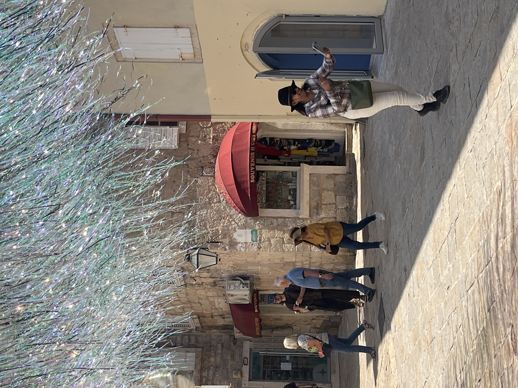 The image captures a bustling street scene in the city of Budva, Montenegro. The street is lined with stone buildings housing souvenir shops and other commercial establishments, each displaying red awnings and shop signs. A group of tourists meanders along the cobbled street, while another person is taking a selfie with a phone. A decorative, shiny, metallic sculpture stands prominently on the left, partially obstructing the view of the buildings. The scene is bathed in sunlight, casting shadows across the street and highlighting the textures of the stone architecture.