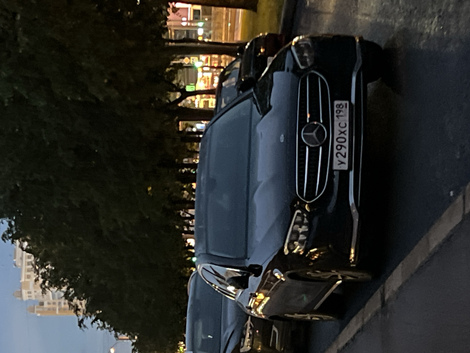 The image shows the front of a black Mercedes-Benz car parked on a street. The car is angled towards the viewer, with its headlights and grille clearly visible. The license plate, which reads "Y290XC 198", is also prominent.
Behind the car, trees line the street, their dark foliage creating a strong contrast with the car's black paint. In the background, a building with multiple floors and windows can be seen, suggesting an urban or suburban setting. The lighting suggests it is either dusk or night, with some artificial lights visible from the building and potentially streetlights. The overall scene is dimly lit, with reflections on the car's surface indicating a damp or wet road. No people are visible in the image. The scene is static, with the car simply parked on the side of the road.