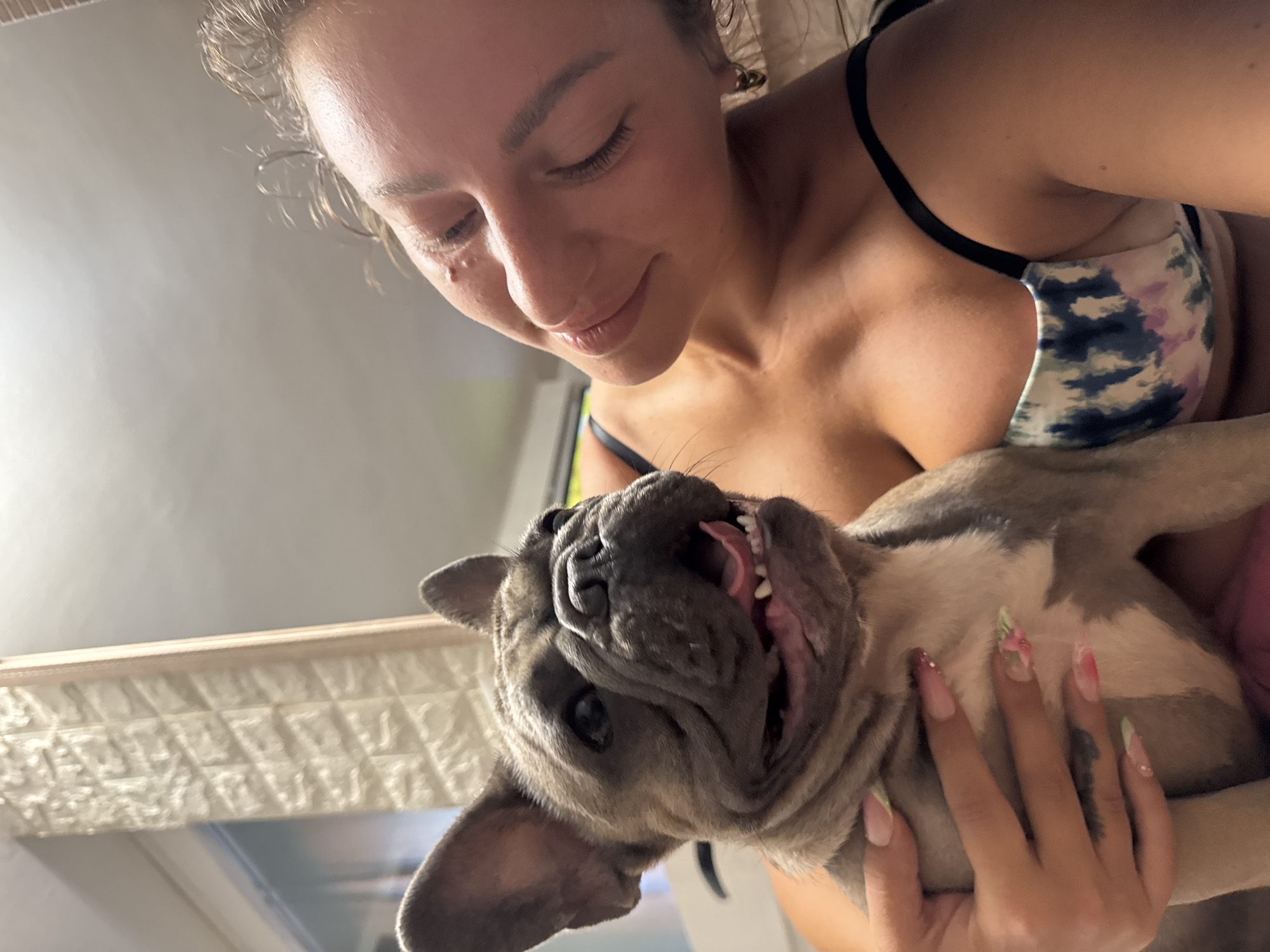 A close-up, indoor shot shows a woman holding a French bulldog puppy. The woman, who appears to be in her twenties, has her eyes closed and is leaning her head towards the puppy. She is wearing a black strap bra with a tie-dye pattern of blue, purple, and white. Her fingernails are long and decorated with colorful designs. The puppy, facing the camera, has its mouth open, revealing its teeth and tongue. Its fur is a mix of gray and tan. The background is blurred, but suggests an indoor setting with textured walls and a hint of natural light coming from a window. The image appears to be taken in San Benedetto del Tronto, Italy.