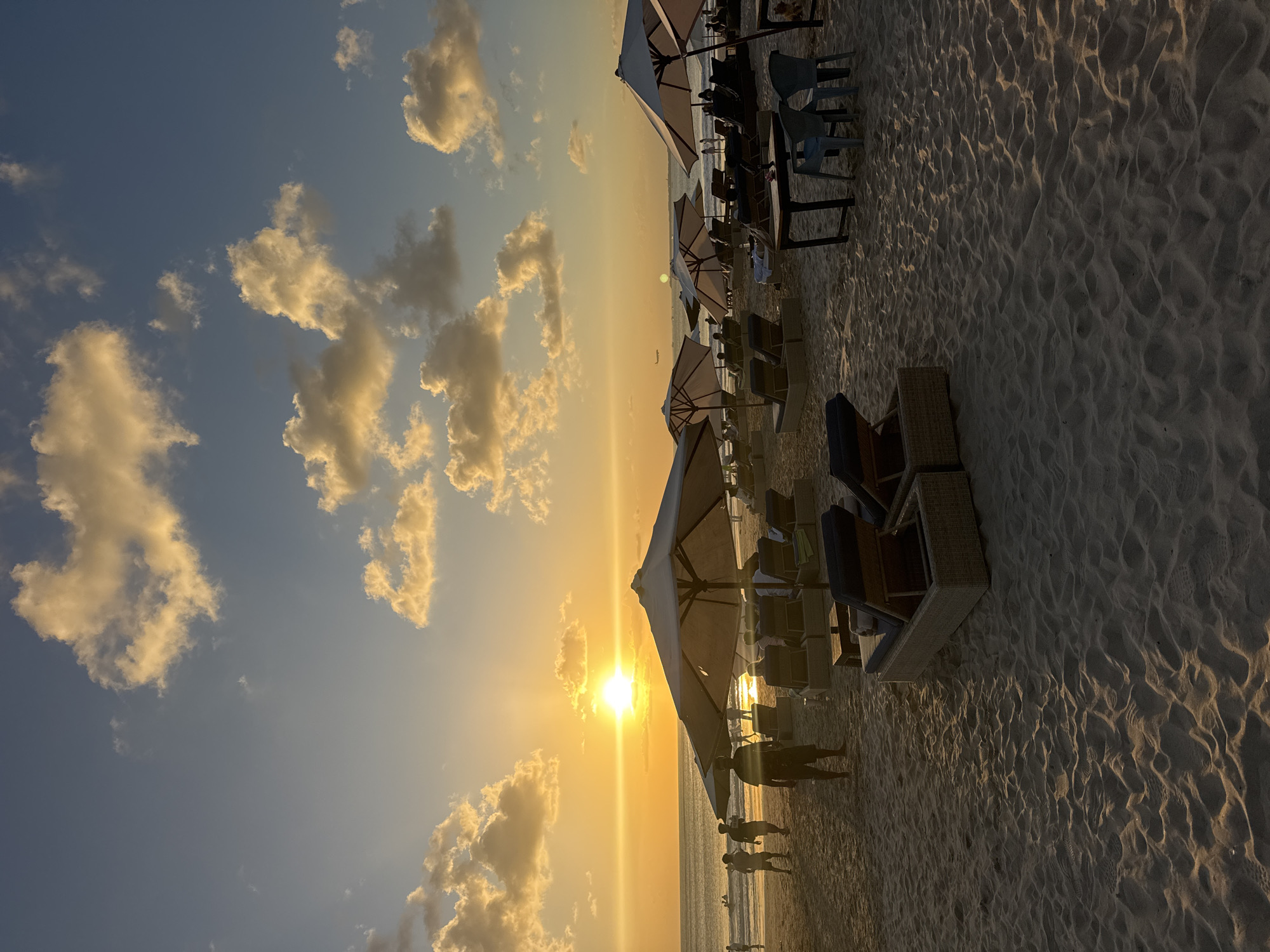 The image depicts a sunset scene on Kuta Beach in Kuta Selatan, Indonesia. The sun is low on the horizon, casting a warm, golden glow across the sky and the sandy beach. Scattered white clouds are illuminated by the setting sun. Several beach umbrellas and lounge chairs are set up on the sand, some occupied by people. In the background, the ocean is visible with small waves, and some individuals are wading or playing in the water. A few people are walking and standing on the beach, and an airplane is visible in the distant sky. The sand shows footprints and ripples from the wind.