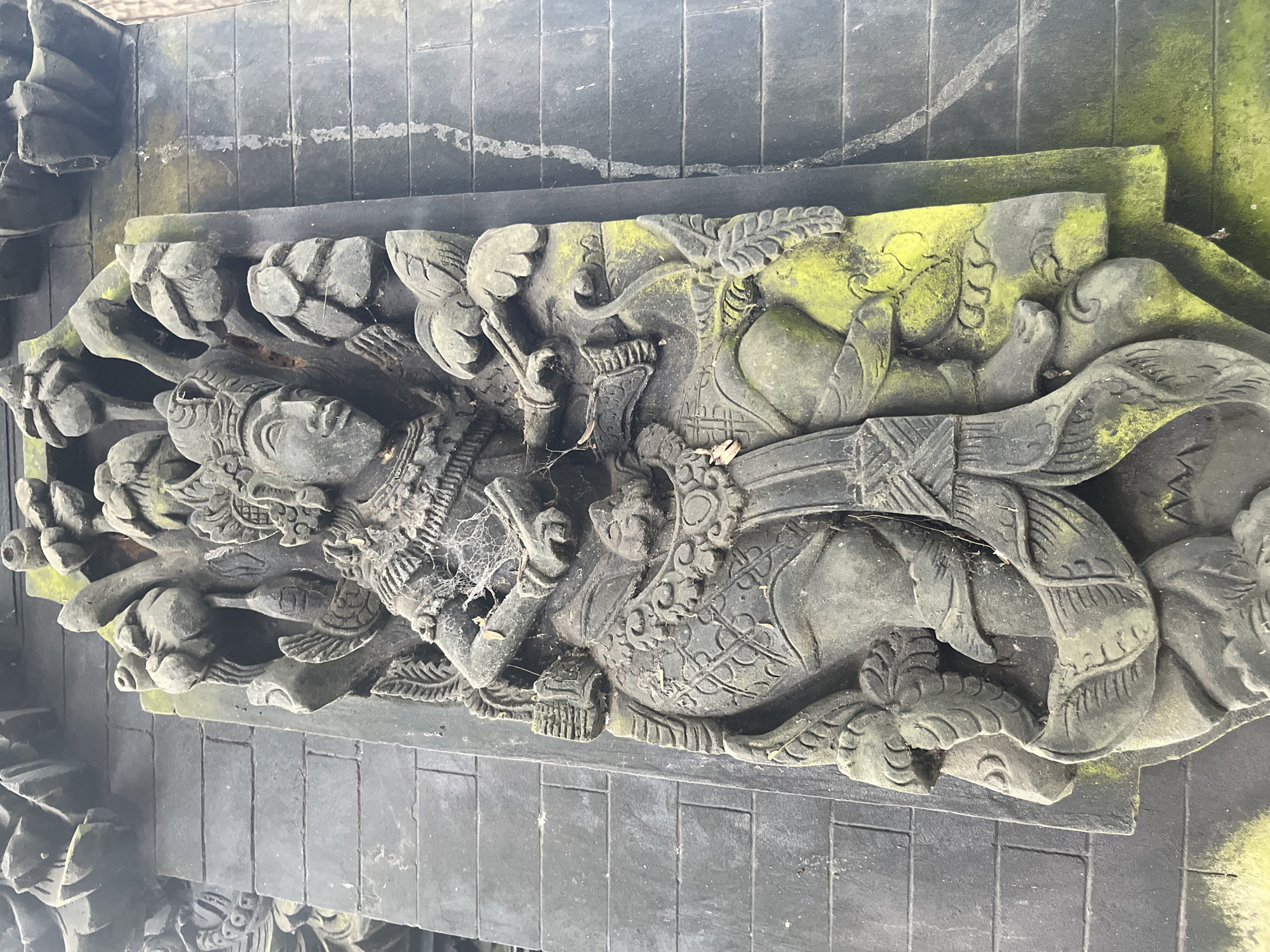 The image displays a close-up, vertically oriented view of a stone relief carving. The carving is mounted on a dark, brick-patterned wall. The relief depicts a human-like figure in a seated position, adorned with ornate headdress and garments. The figure is surrounded by carved foliage and appears to be holding an object in its hands. The stone surface of the carving exhibits significant weathering, with patches of green moss or algae growing on it, particularly on the lower half of the figure and the surrounding elements. Some areas of the carving are covered in fine cobwebs. The background wall consists of evenly spaced, rectangular dark bricks. The overall impression is of an ancient, weathered piece of art integrated into a structural element. The location is identified as Kuta Selatan, Indonesia.