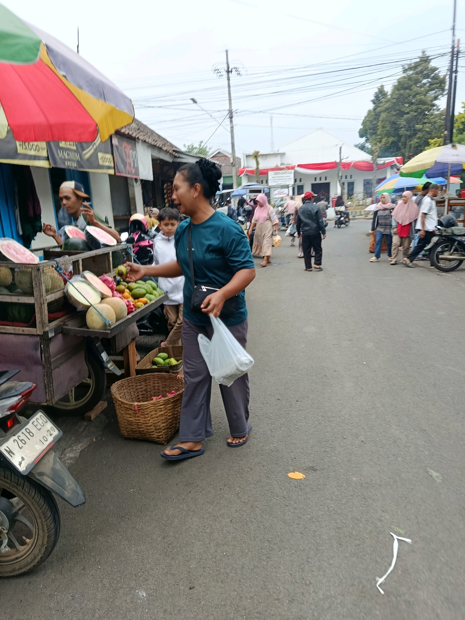 The image depicts a street scene in Bululawang, Indonesia, likely a market or roadside stall. A woman in a teal shirt and grey pants stands in the foreground, holding a white plastic bag and reaching towards a fruit stand. The fruit stand, mounted on a cart, displays various fruits including melons, papayas, and what appear to be mangoes or avocados. Behind the woman, a young boy in a white shirt looks towards the fruit stand. To the left of the fruit stand, a vendor wearing a cap is visible, appearing to serve a customer. Further in the background, other people are walking or standing, some wearing hijabs, and motorcycles are parked or being ridden on the street. The environment includes a paved road, several buildings with awnings and signage, and overhead power lines. A basket filled with fruits is placed on the ground next to the fruit stand. A motorcycle license plate with "N 2618 ECO" is visible on the left side of the image. The sky is overcast.