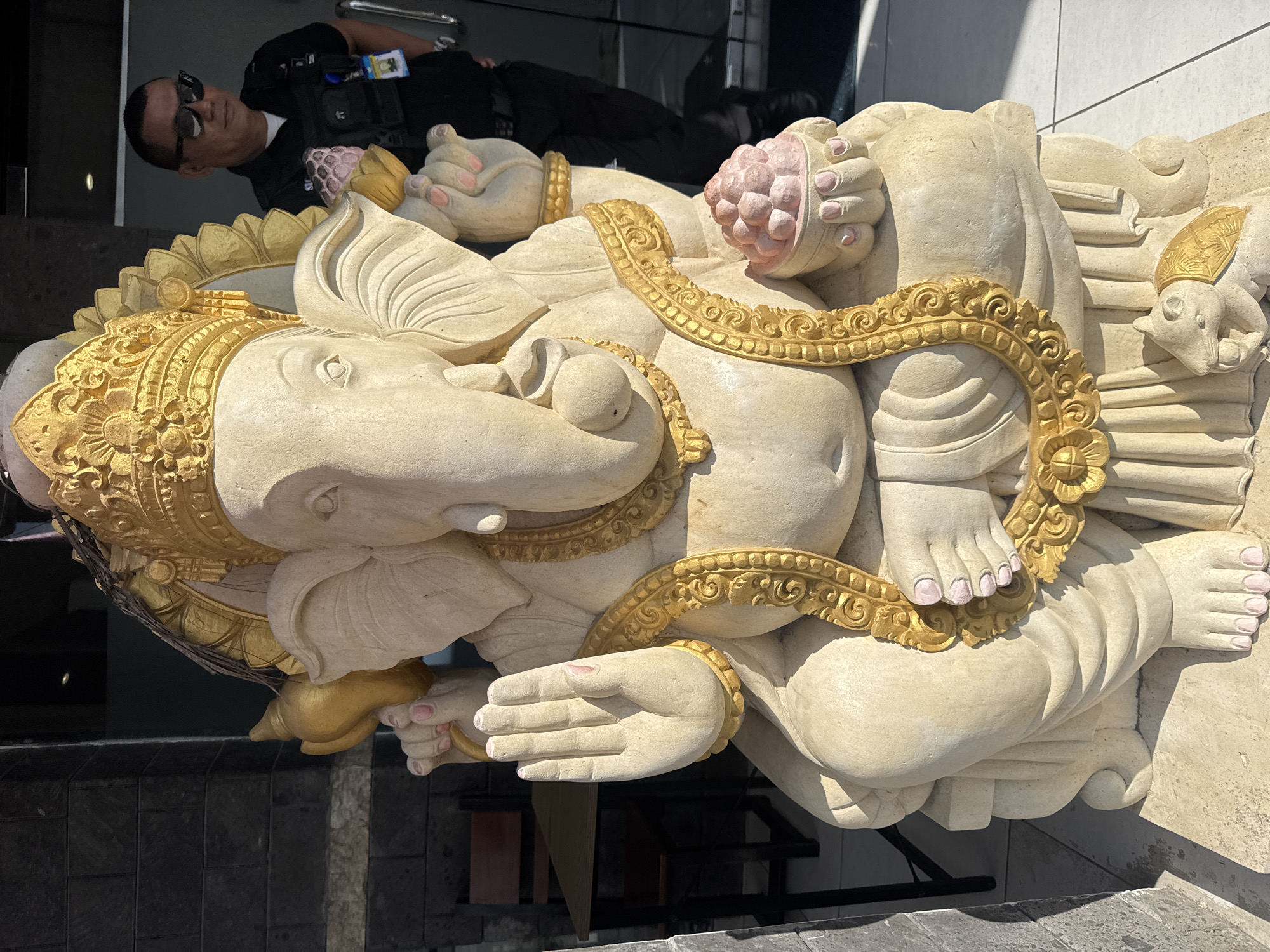 A statue of Ganesha sits on a sidewalk in Kuta Selatan, Indonesia. The light-colored stone sculpture of the Hindu deity is adorned with gold ornamentation. Ganesha is seated cross-legged, holding what appears to be a pomegranate in one hand and a conch shell in another. A man in a dark uniform and sunglasses is standing in the background.