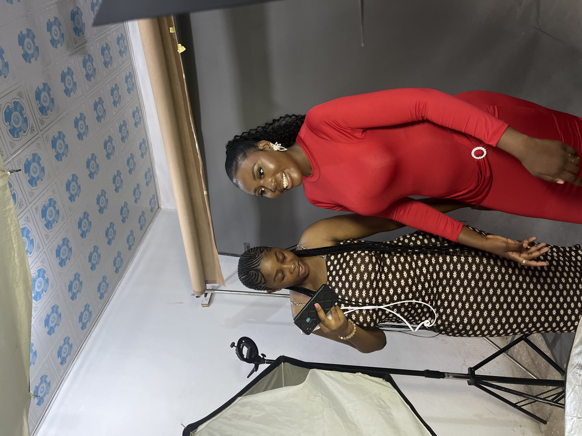 Two young Black women are standing in what appears to be a studio setting. The woman on the left is wearing a brown and white patterned dress and is holding a black smartphone. She has her hair braided in cornrows. The woman on the right is wearing a long-sleeved red dress with a circular sparkly embellishment at the waist. She is smiling and looking to her left. The background consists of a grey backdrop and a light-colored roll of paper or fabric. To the left of the frame, there is a photography light setup with a large diffuser and a stand. The ceiling has a patterned tin tile design with blue floral motifs. There is no indication that this scene is located in Gura Top, Nigeria, or any specific geographical location.