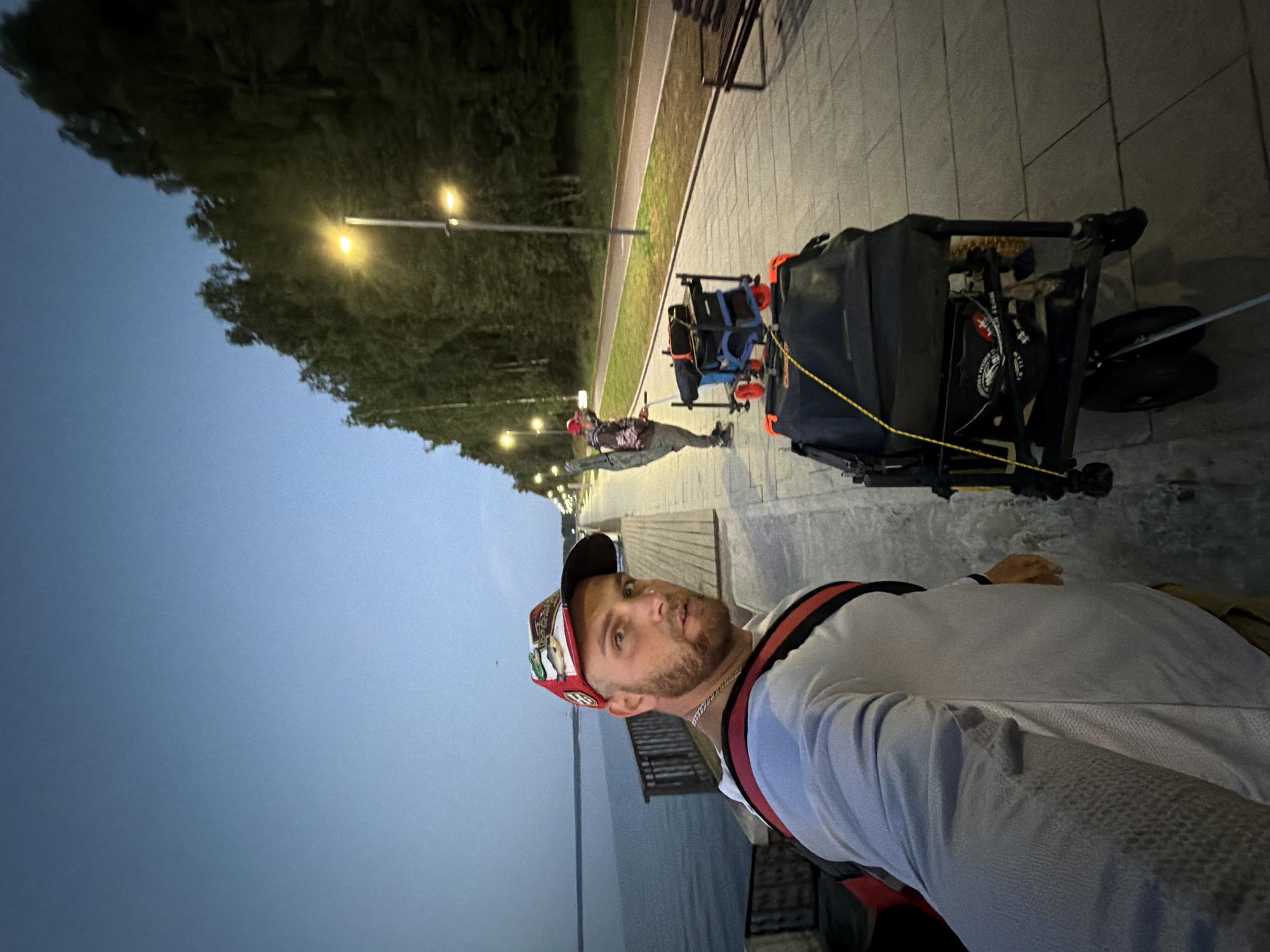 A man in a baseball cap and long-sleeved shirt is taking a selfie. He is standing on a paved walkway next to a body of water, possibly a river or lake. Behind him, another person is visible, pulling a cart with fishing equipment. The walkway is lined with trees and streetlights, suggesting an urban park or waterfront area in the city of Chaikovsky, Russia. The sky is dark, indicating it is either early morning or evening.