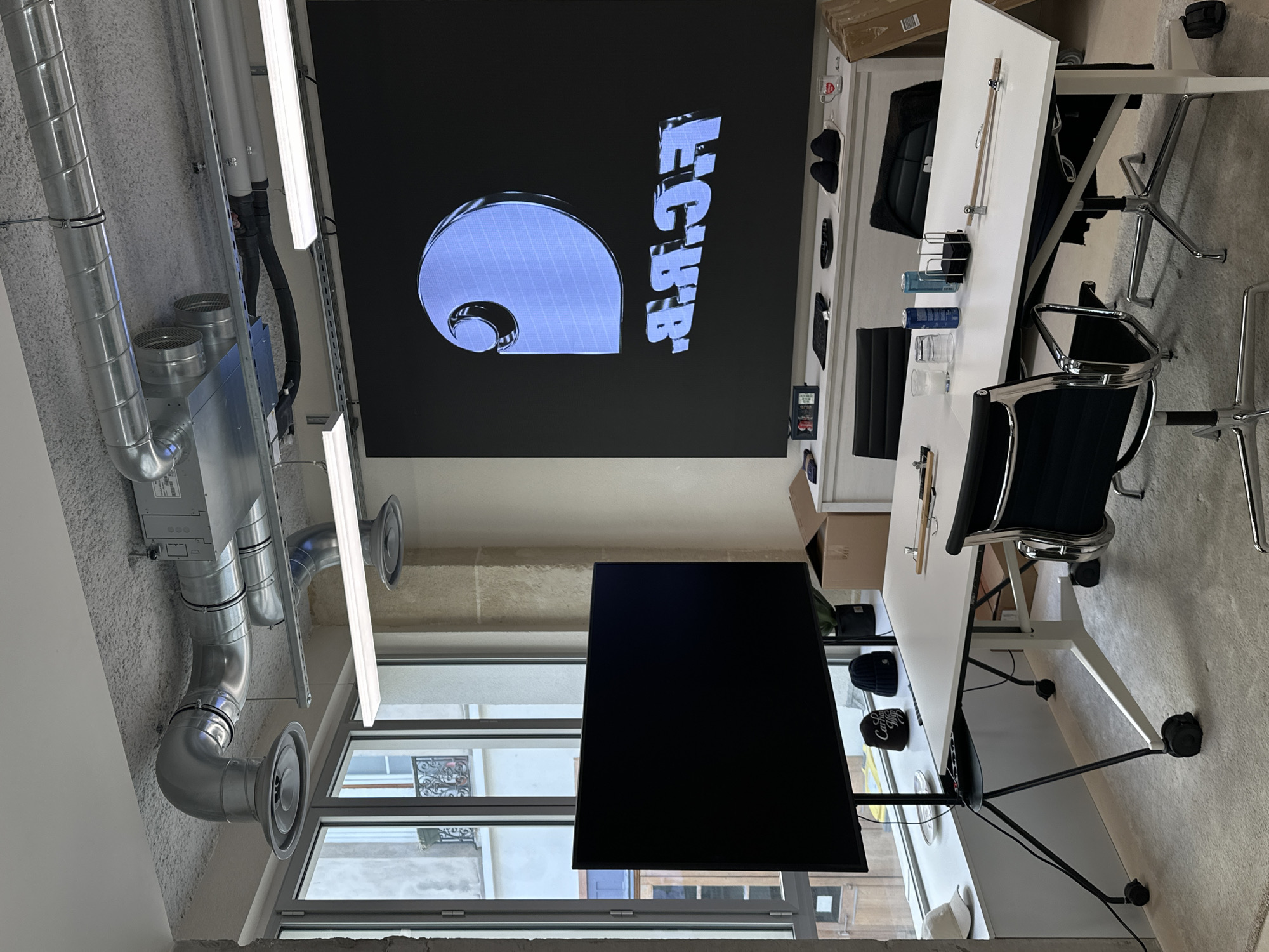 The image shows an office space with a large screen displaying a logo, likely inside a building in Paris, France, as suggested by the architectural style of the building visible through the windows.
The office setup includes:
* **Screen:** A large screen displaying the Carhartt logo in blue and black.
* **Ventilation System:** A visible network of metal ventilation ducts and fans running along the ceiling.
* **Lighting:** Fluorescent tube lights installed along the ceiling.
* **Window:** A large window. The exterior view includes a building facade with wrought-iron balconies, which are characteristic of Parisian architecture.
* **Furniture:** A white table with chairs.
* **Objects:** There are various objects on the table and on shelves, including clothing, boxes, and other office supplies.
* **Wall Décor:** Items on the wall include hats, small pictures, and possibly mugs.
The scene appears to be a modern office with a mix of industrial and contemporary elements. The overall impression is that of a functional workspace with branded elements.