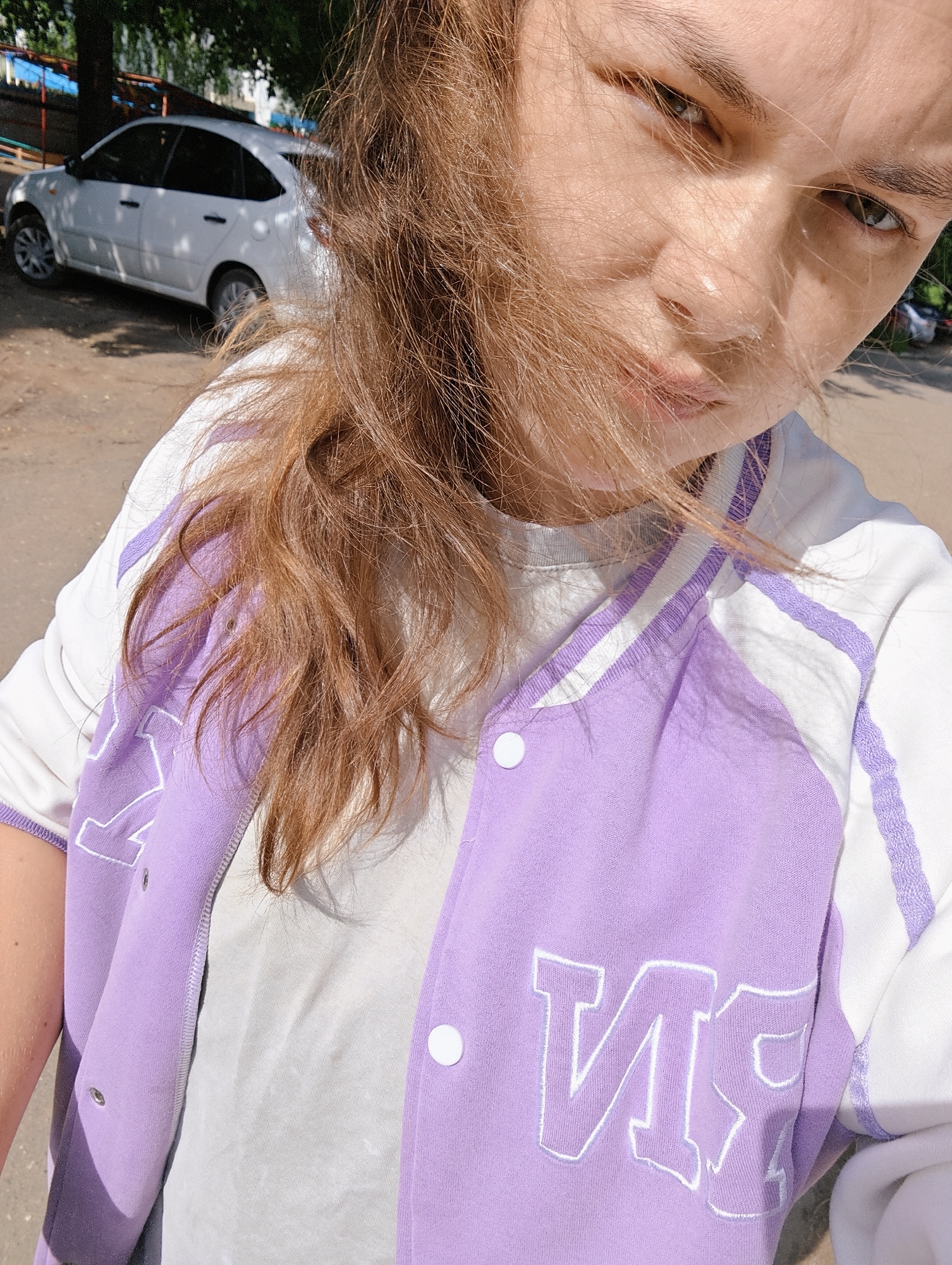 The media file presents a close-up, selfie-style portrait of a young person, likely female, outdoors on a bright, sunny day in Chaikovsky, Russia.
The primary subject is looking directly at the camera with a neutral expression. Their light brown or auburn hair is partially blown across their face, creating a dynamic effect. They are wearing a light-colored (possibly white or light grey) t-shirt, over which they have a distinctive varsity-style jacket. The jacket is predominantly lavender (light purple) with white sleeves, white snap buttons, and white striped trim on the collar and shoulders.
Visible text on the jacket, embroidered in bold white letters, appears reversed due to the selfie camera. On the left side of the jacket (viewer's left), the Cyrillic letters "ИЯ" are clearly visible. A partial, reversed letter, possibly a "V", is seen on the right side.
In the blurred background to the left, a white sedan car is parked under some trees, suggesting an urban or suburban setting. The ground appears to be a mix of dirt and paved surfaces. The strong sunlight illuminates the scene, indicating it is daytime with clear weather. The overall impression is an candid outdoor self-portrait capturing a moment in bright conditions.