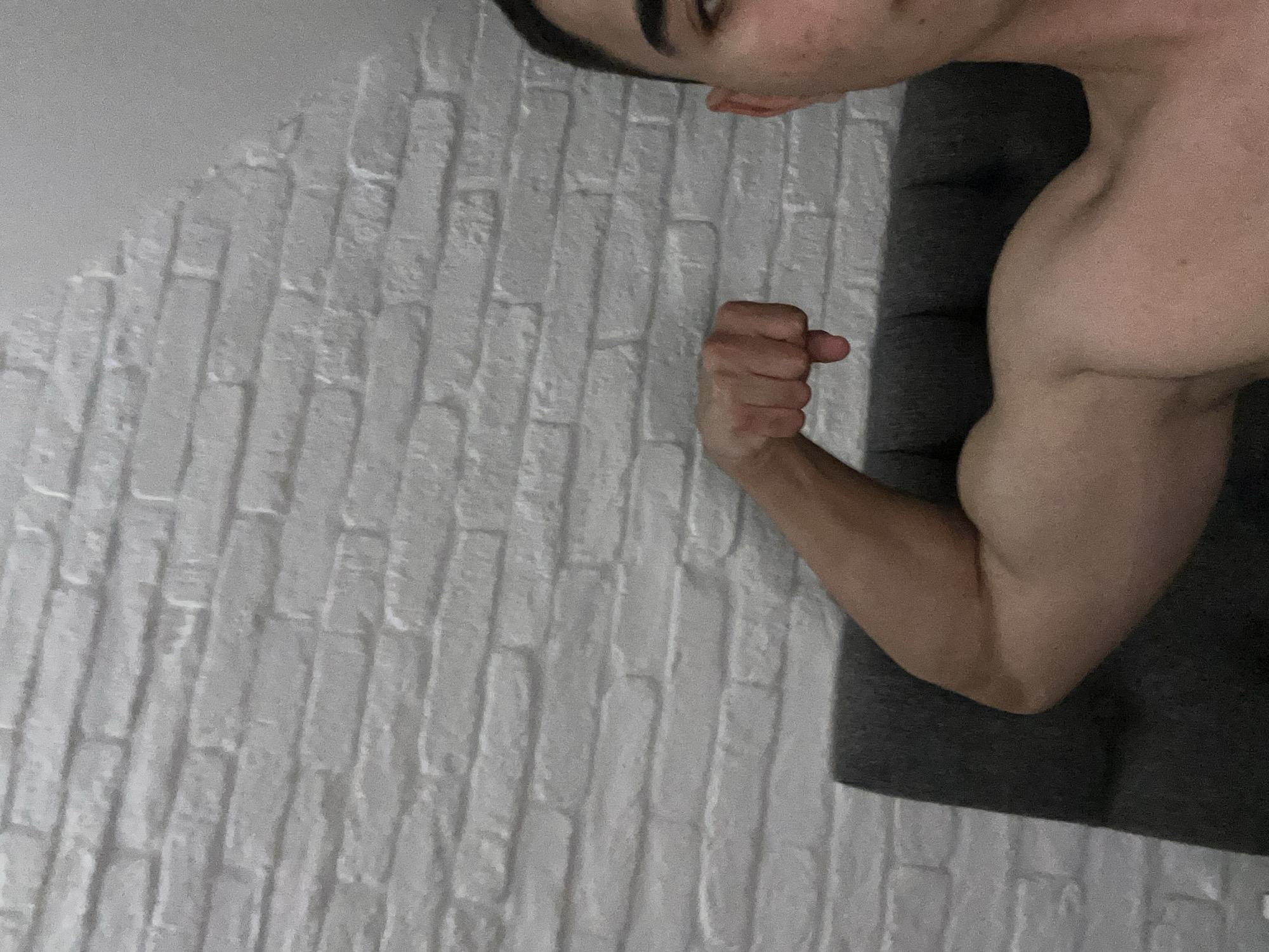 A person is flexing their right arm. The background consists of a white brick wall and a grey cushioned surface. The subject's head is partially visible in the upper portion of the image. The location is not discernible from the image.