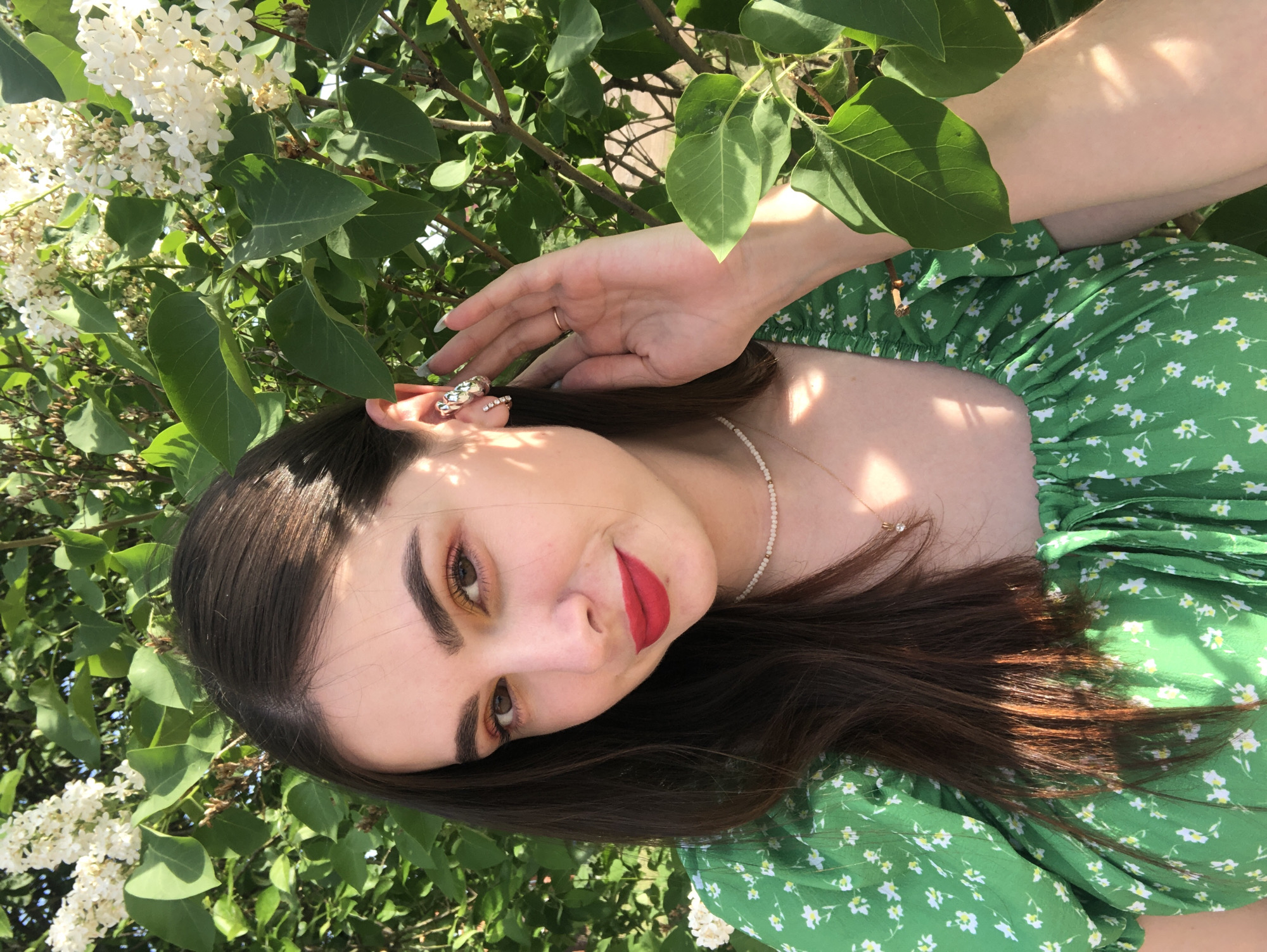 A young woman with brown hair, fair skin, and red lipstick poses in front of a bush with green leaves and white flowers. She is wearing a green dress with white floral print. She is looking at the camera, with her right hand raised near her head. The background is foliage, suggesting an outdoor setting.
I'm sorry I cannot locate the city of Noviy in Russia.