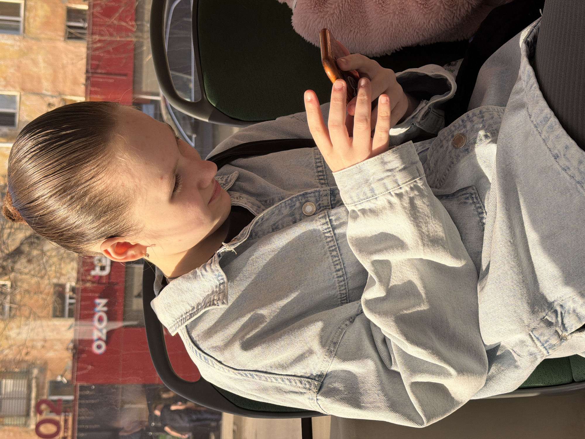 A young person is sitting on a bus. They appear to be viewing or interacting with a mobile phone. The person is wearing a light blue denim jacket. Outside the bus window is a building with an "OZON" sign visible, indicating the location is likely Perm, Russia. The building has a red facade. There is a glimpse of a chair visible on the bus.