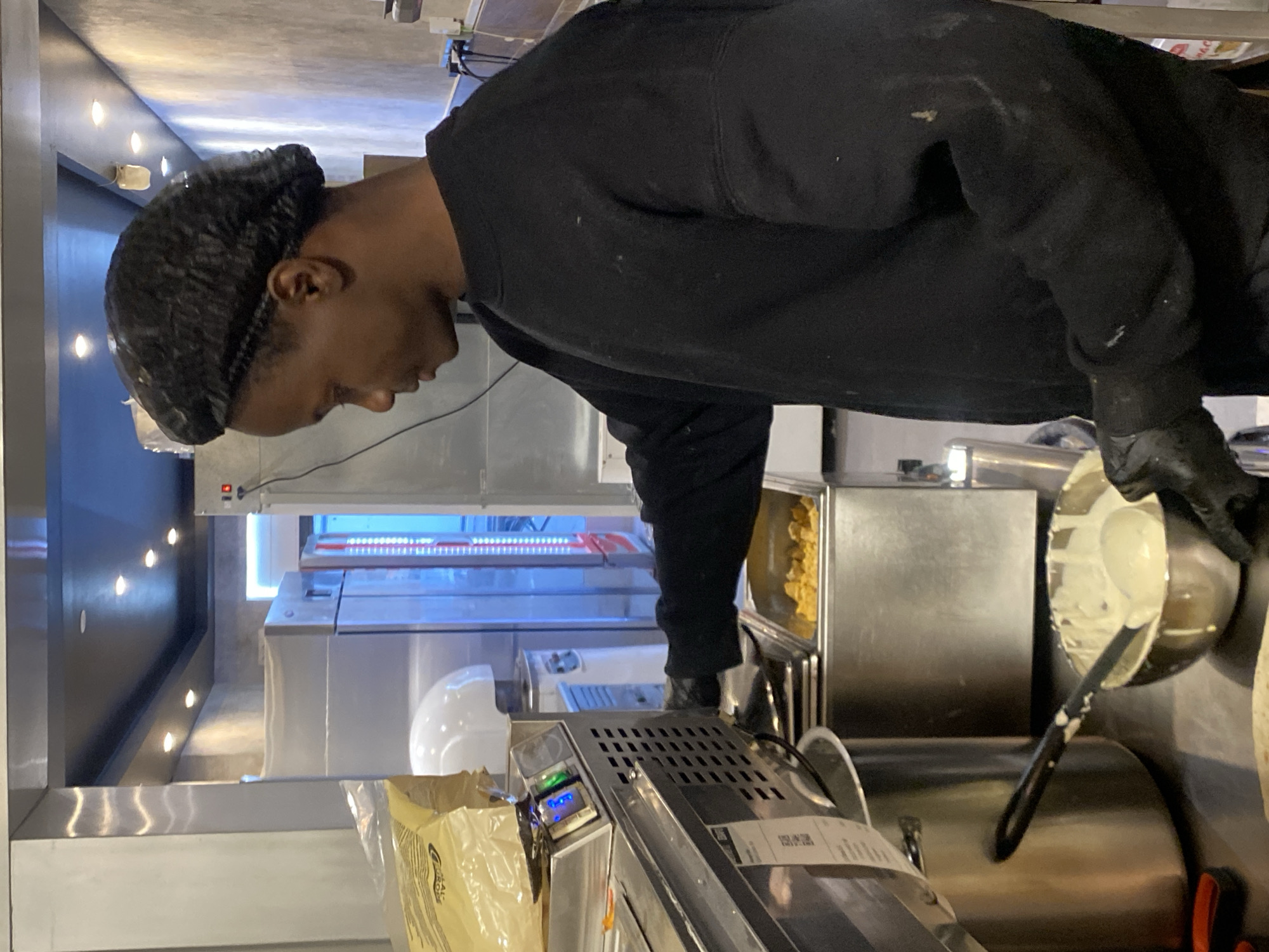 A person wearing a black hair net, black sweatshirt, and black gloves is working in a commercial kitchen. The kitchen appears to be made of stainless steel, with visible appliances, containers, and tools. The location is Schiltigheim, France. The person is holding a metal bowl containing a light-colored substance.