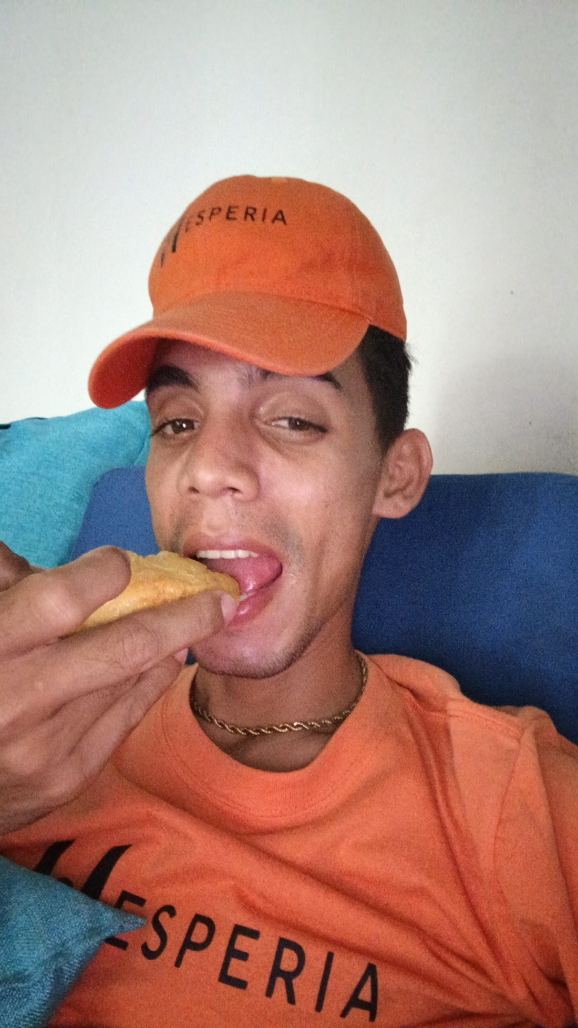 A young man is eating a handheld food item, possibly an empanada, while seated on a blue cushion or couch. He is wearing an orange t-shirt and a matching orange baseball cap, both emblazoned with the word "ESPERIA" in black lettering. He also wears a gold chain around his neck. The background is a plain white wall. The image appears to be a selfie. There is no evidence to confirm that the photograph was taken in Playa El Agua, Venezuela, as this information is not visually discernible.