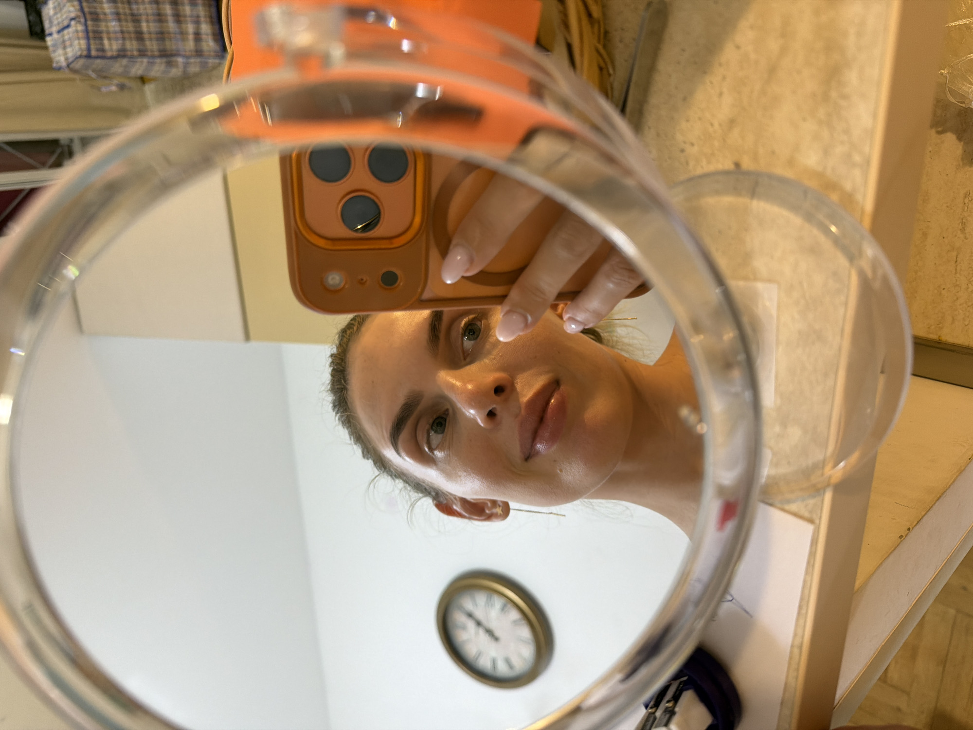 A woman is taking a selfie in a circular mirror. She is holding an orange iPhone in her right hand, with her fingers extended and nails painted a light pink. Her left hand is also visible. Her face is partially visible in the mirror, with her eyes looking slightly to the left. She has long, light brown hair pulled back. She is wearing a light-colored top.
In the reflection of the mirror, a round wall clock with a gold frame is visible on a white wall. The clock has black Roman numerals and hands.
The background appears to be an indoor setting, possibly a room with a desk or counter where the mirror is placed. There are some items on the counter, including a dark blue item and some white paper. In the far background, a plaid duffel bag is visible.
The scene is lit with natural light, casting a soft glow. The image is taken from a low angle, looking up at the woman and the mirror. There is no indication of the city of Budva, Montenegro in the image itself.