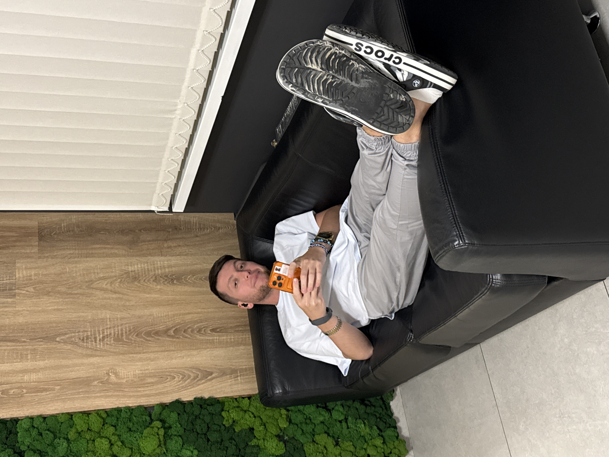 A man reclines on a black leather couch, holding an orange cell phone and facing the camera. He wears a white t-shirt and gray sweatpants, and his feet, clad in dirty Crocs, are propped on the couch back. A window with blinds is visible behind the couch. The floor is a light wood grain, transitioning to gray tile in the lower right corner. Below the wood floor, there is a patch of bright green moss. While the location within Moscow, Russia, cannot be definitively confirmed from the image, the image depicts an indoor setting with modern decor.