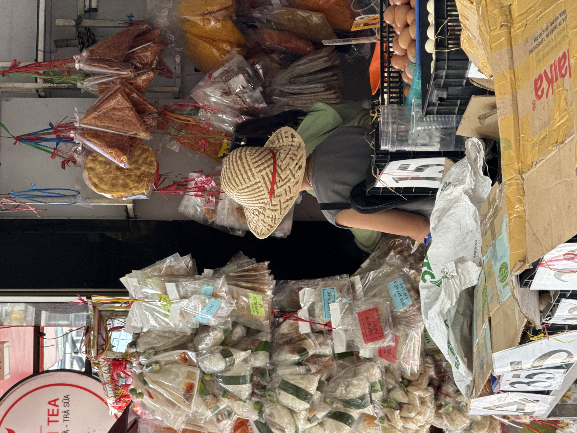 The image shows a street market stall in Nha Trang, Vietnam. A person wearing a straw hat is standing behind a display of various goods. The stall is filled with bags of food items, some of which are hanging from the ceiling. There are also crates of eggs visible in the foreground. The overall impression is one of a bustling market with a wide variety of products for sale.