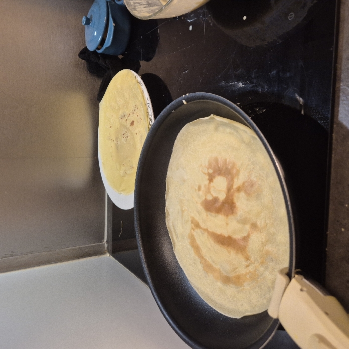 A top-down shot showcases a kitchen scene. A pancake browns in a skillet with a beige handle. To the left, a cooked pancake rests on a white plate. Above the skillet, a blue teapot and a bowl of batter sit on a black stovetop. The scene includes stainless steel surfaces, a black potholder, and a glimpse of a textured countertop.