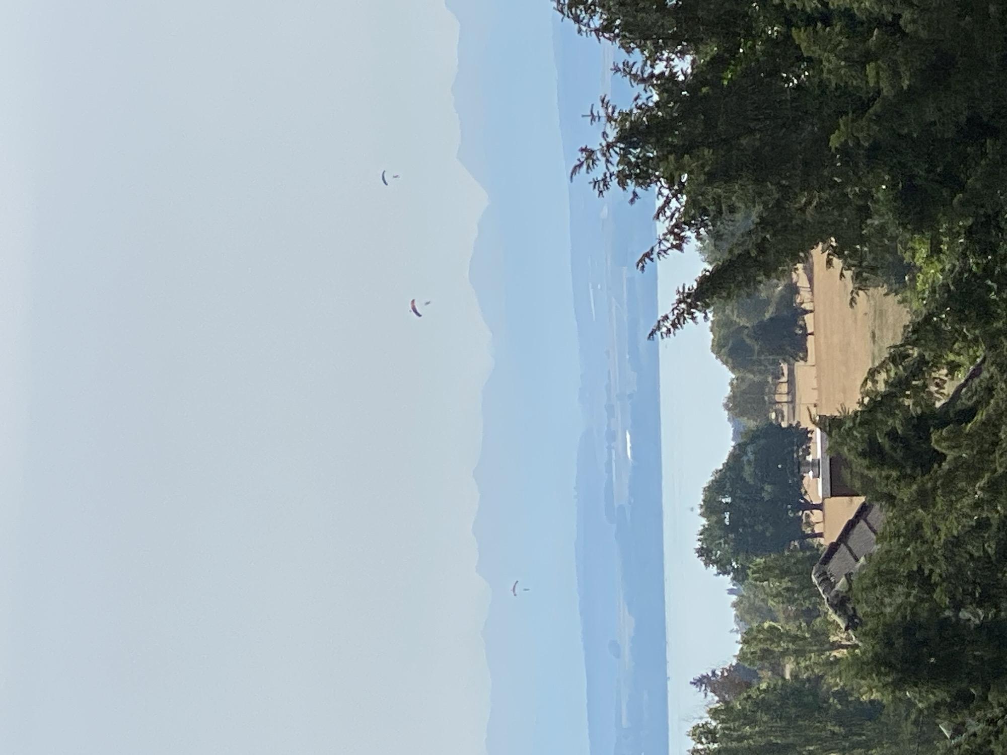 The image captures a panoramic view of a lake and distant mountains under a hazy sky. In the foreground, lush green trees partially obscure a rustic house with a pitched roof and a chimney. Beyond the trees, a vast body of water stretches out, reflecting the muted light of the sky. The middle ground features a dry, grassy field dotted with trees, suggesting a rural or park-like setting. Further in the distance, the soft blue outlines of mountains create a layered effect against the horizon. Three paragliders are visible in the sky, appearing as small, colorful dots against the vast expanse. One paraglider is further to the left and slightly lower than the other two, which are closer together in the center of the frame. The overall atmosphere is serene and expansive.
