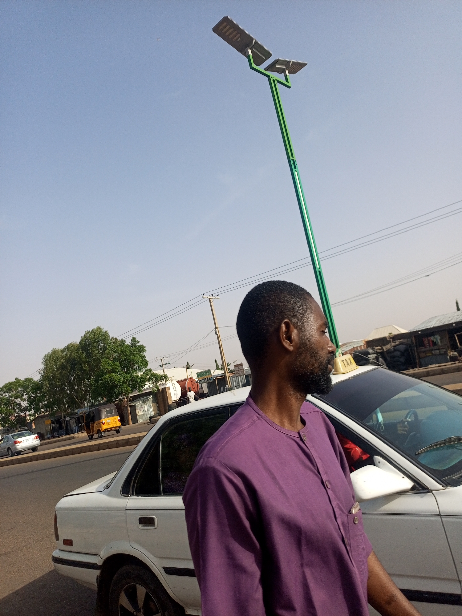The image captures a street scene in Gombe, Nigeria. A man wearing a purple traditional shirt stands next to a white taxi. The taxi has a "TAX" sign on top. In the background, other vehicles are visible, including a yellow tricycle taxi (keke). A tall, green solar-powered street lamp stands adjacent to the man. Buildings are visible in the background, as are overhead power lines. The sky is pale blue.