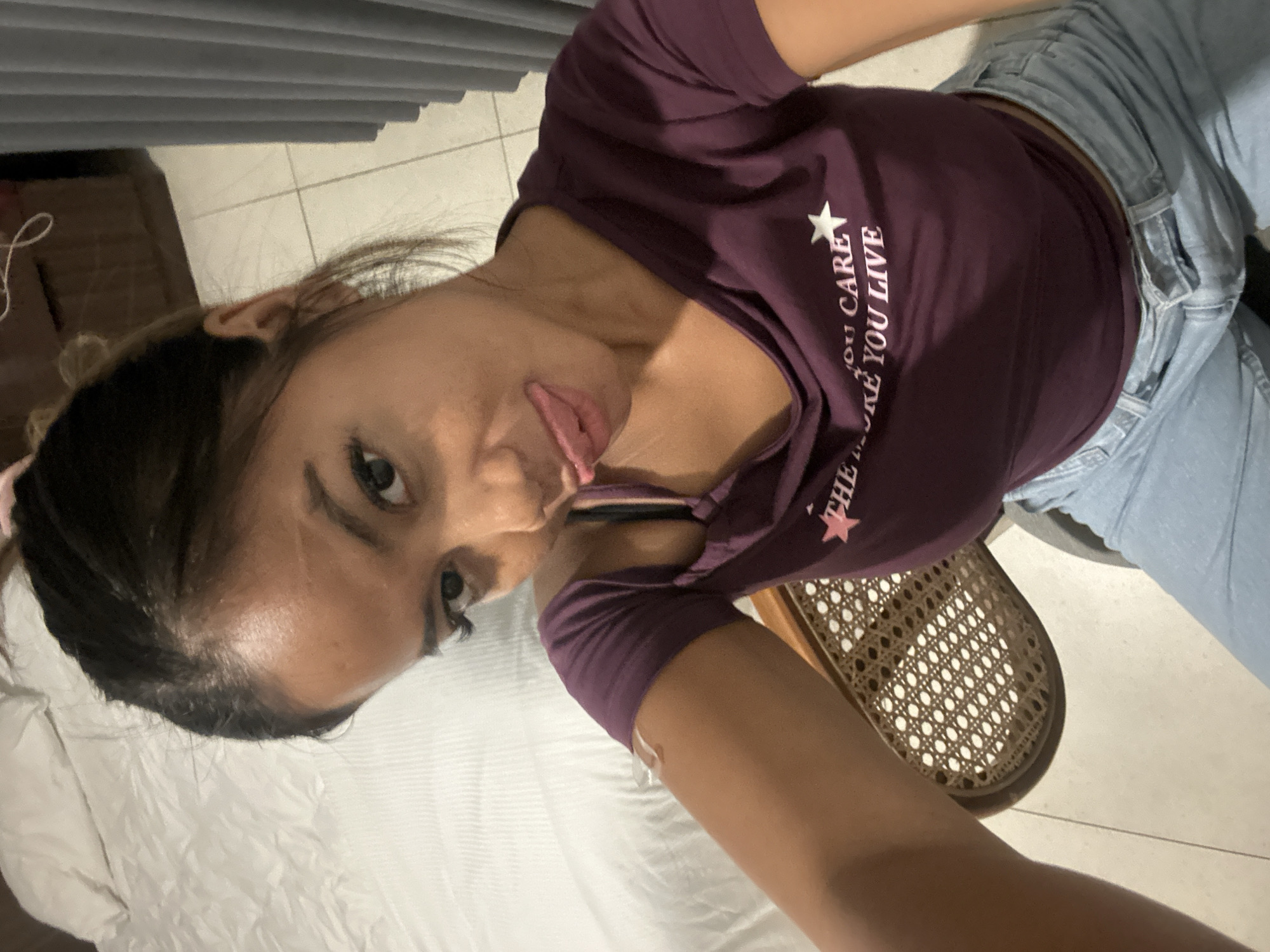 A young woman with dark hair pulled back and a casual updo, wearing a maroon t-shirt and light blue jeans, is pictured in what appears to be a bedroom. She is sticking her tongue out playfully at the camera. A white bandage is visible on her upper arm. The room contains a bed with white linens, a cane-backed chair, and a grey curtain in the background. The floor is tiled. This image was taken in North Kuta, Indonesia.