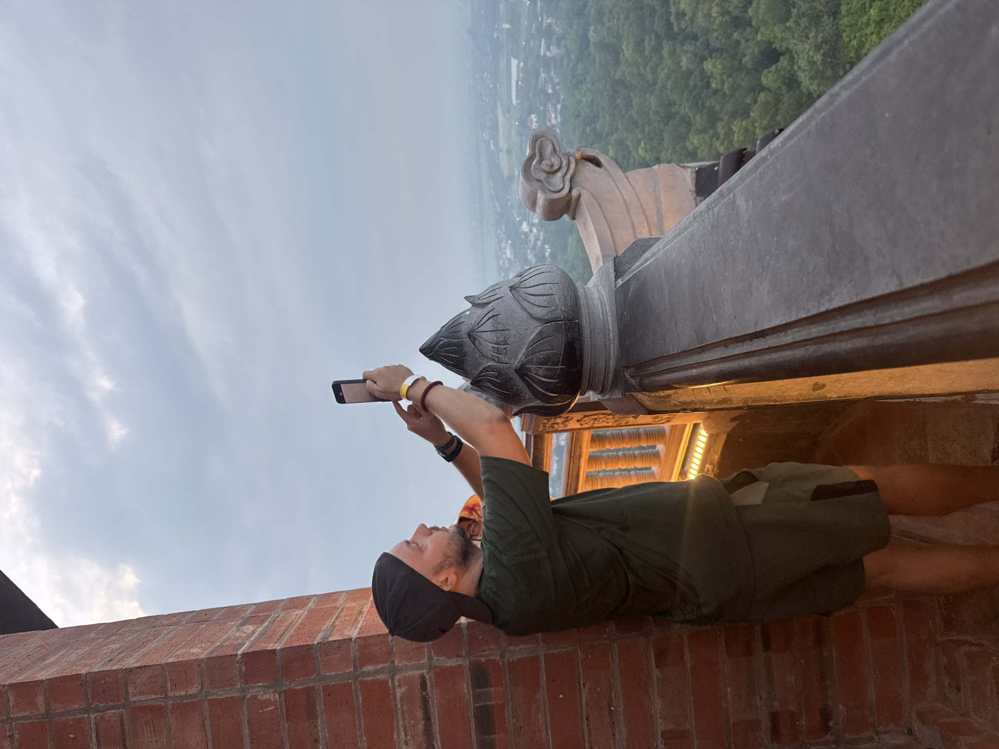 A man is taking a photo with his phone on a balcony overlooking a misty landscape. He is wearing a green t-shirt, shorts, and a black baseball cap. He is standing next to a brick wall and a railing with decorative carvings. The background shows a city with trees and hills in the distance. The lighting suggests it is dusk or dawn. The location is likely a viewpoint in Tây Hoa Lư Ward, Vietnam, given the architectural style and the misty, tropical landscape.