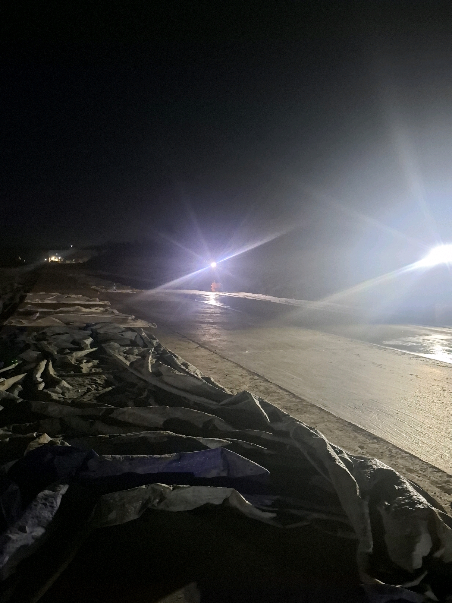 A nighttime photograph captures a construction scene in Karangtengah, Indonesia. The foreground is dominated by large, grey tarps piled on the ground, some partially illuminated by artificial light. A paved surface, likely a road or construction area, stretches into the background. Bright lights, possibly from construction equipment, create strong glare and lens flare effects, illuminating the wet or reflective surface of the paved area. In the distance, a few figures can be seen, and there are hints of structures or other elements of the urban or construction environment. The overall impression is of a worksite operating after dark.