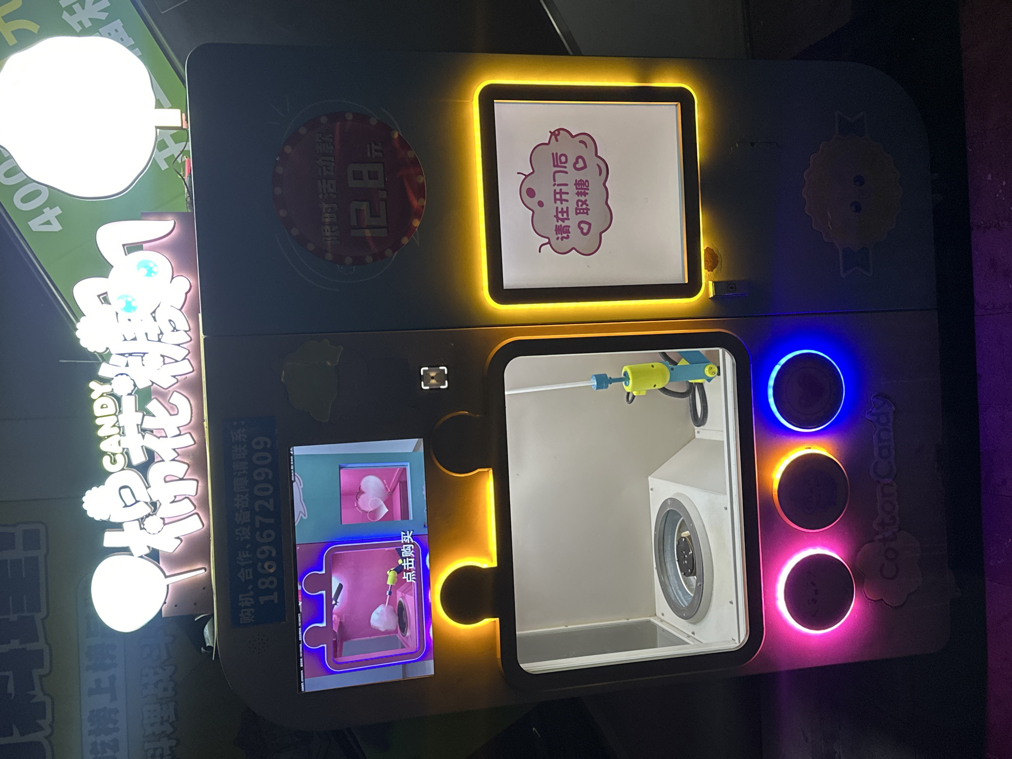 Here is a detailed description of the image:
**Scene:** The image showcases a cotton candy vending machine, likely located in the city of Chongqing High-tech Zone, China, based on the Chinese characters present on the machine.
**Details of the Machine:**
* **Exterior:** The machine has a box-like design, illuminated with colorful lights, including blue, orange, and pink. The exterior is decorated with cute cartoon-like images.
* **Signage:** There's a prominent sign on the left that reads "科理战斗 CANDY," which could be the machine's brand or marketing phrase. Beneath this is the phrase "吃榜上榜品." A phone number, "18696720909," is listed, likely for inquiries related to purchase, cooperation, or equipment malfunction. The machine also has the text "Cotton Candy."
* **Displays:** The machine has several displays. One shows a promotional price of "12.8 元". Another has the text "请在开门后○取糖," which means "Please take the cotton candy after opening the door". One displays a demo of how the cotton candy is made.
* **Internal Components:** Visible through a glass-fronted compartment is a cotton candy making apparatus, including what appears to be a heating element and a dispensing tool.
* **Buttons:** The machine has three large, circular buttons with colored lights surrounding them.
**Overall Impression:** The machine appears to be a modern, attractive vending machine designed to appeal to customers with its bright lights, cartoonish decorations, and clear display of the cotton candy-making process.