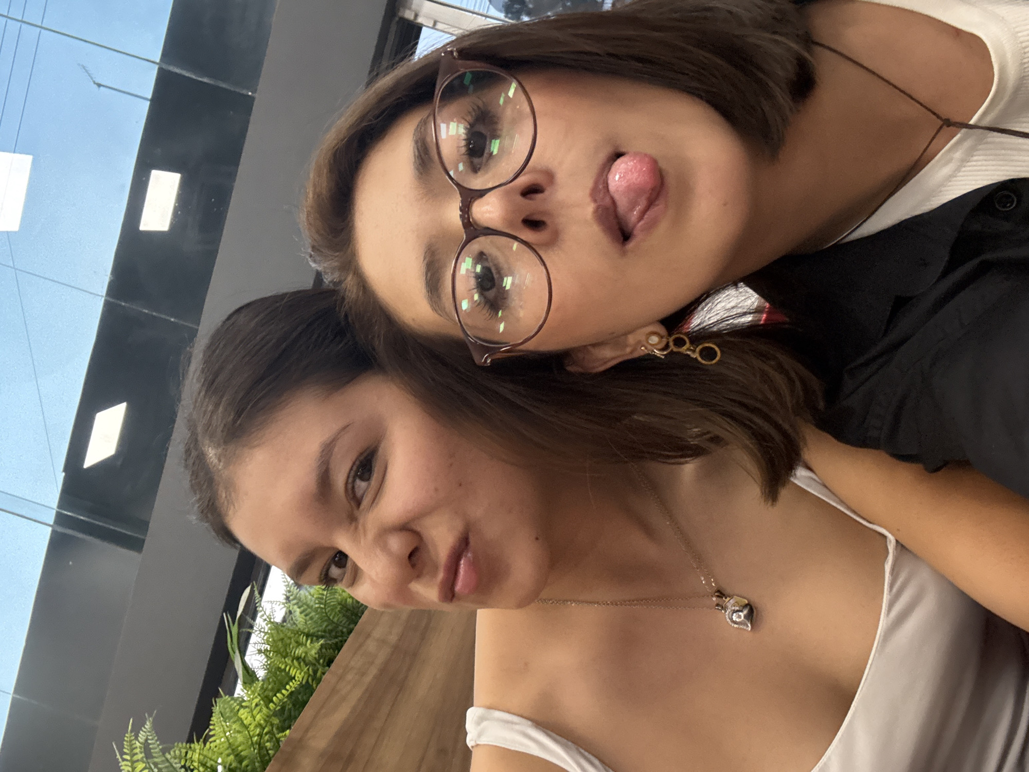 Two young women are posing closely together for a selfie. The woman on top is wearing round glasses and has her tongue out. She is wearing earrings and a black shirt over a white top. The woman below is wearing a white top and a necklace. Green plants are visible on the left side, and there is a window behind them. The location appears to be indoors, possibly a restaurant or cafe in San Andrés Cholula, Mexico.